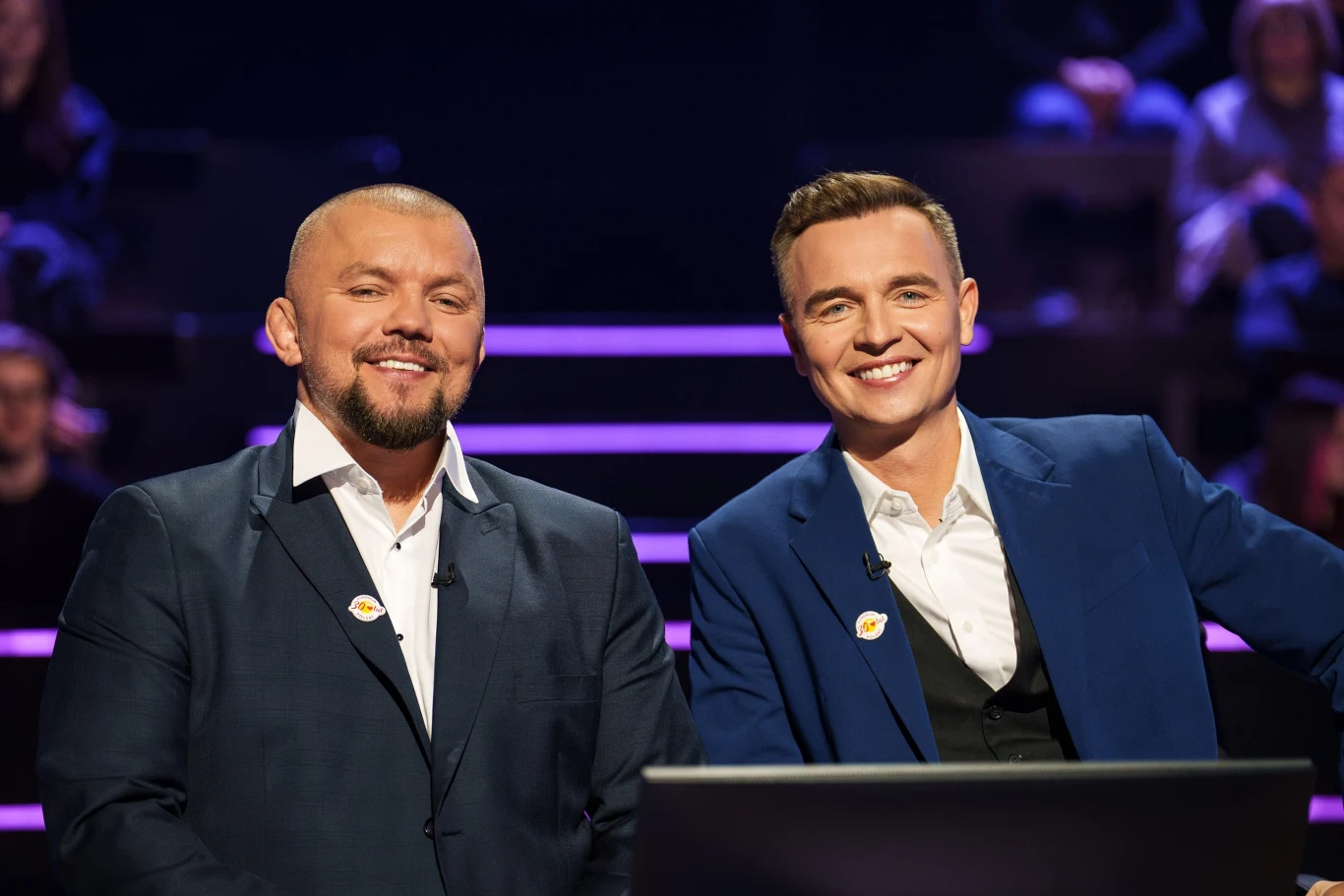 Dwóch elegancko ubranych mężczyzn w garniturach siedzi obok siebie, uśmiechając się w stronę aparatu; w tle widoczne są rzędy siedzących ludzi oraz neonowe, poziome światła podkreślające atmosferę studia telewizyjnego.