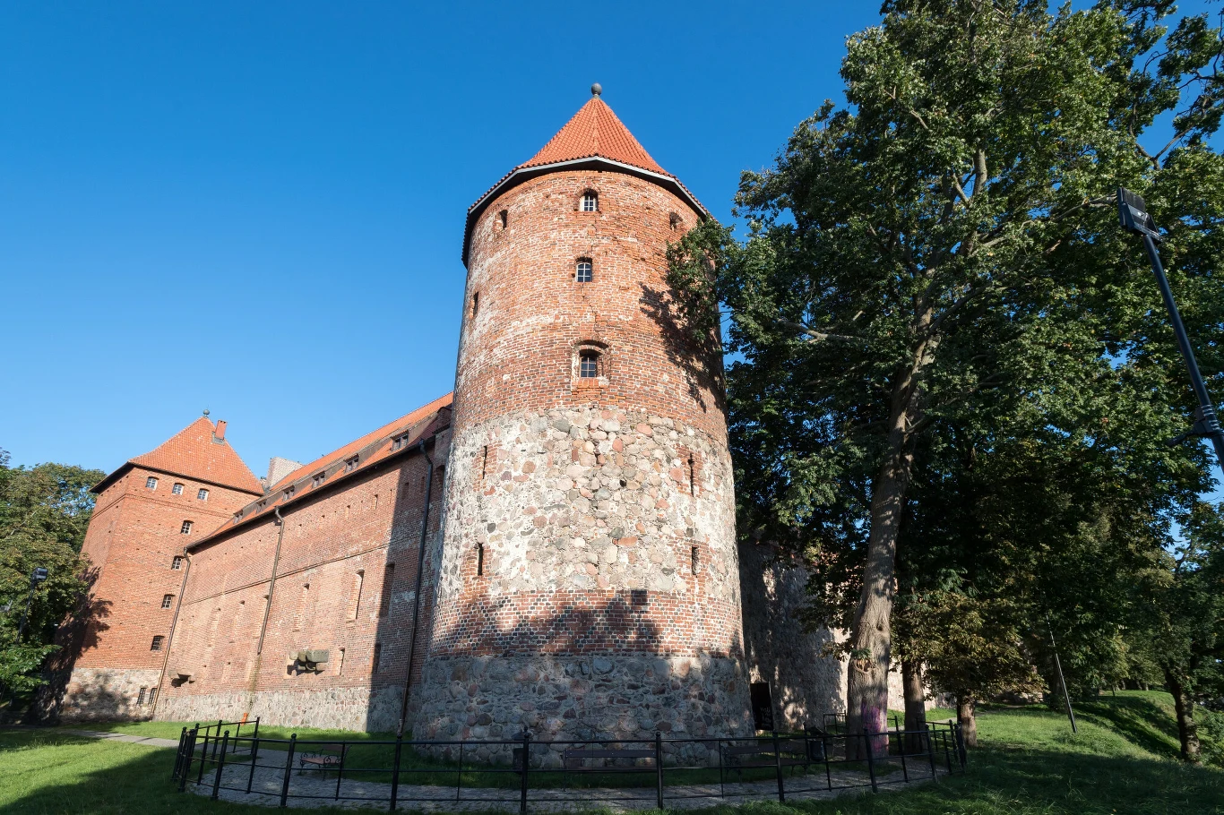 Ceglany zamek z okrągłą wieżą otoczoną kamiennym fundamentem, przykrytą czerwoną dachówką, w otoczeniu drzew i żelaznego ogrodzenia na tle nieba bez chmur.