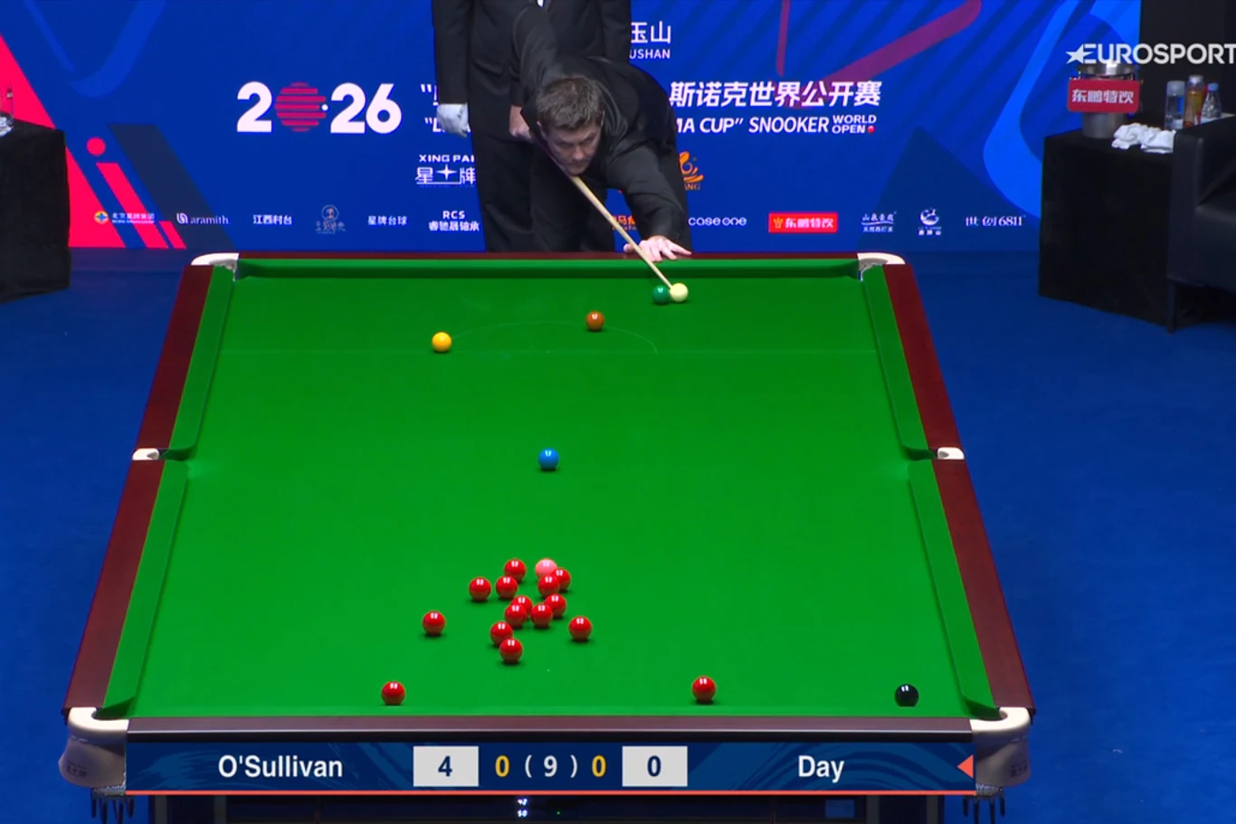zawodnik wykonuje uderzenie kijem bilardowym na stole snookerowym, na którym większość czerwonych bil skoncentrowana jest w centralnej części stołu, wynik meczu widoczny na elektronicznym wyświetlaczu pokazuje prowadzenie O’Sullivana nad Dayem, w tle w...