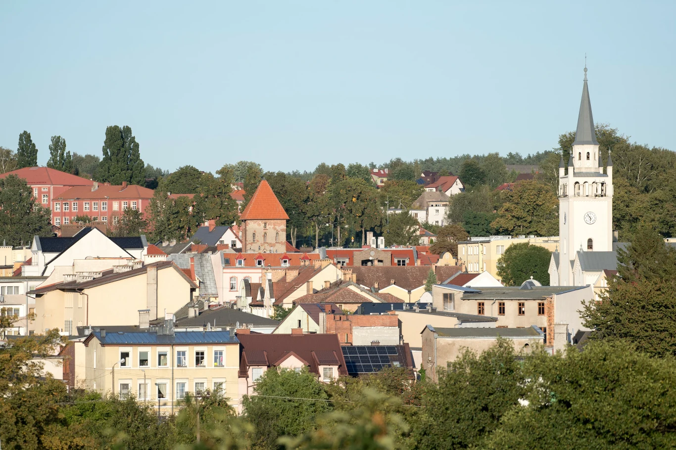 Panorama miasteczka z widocznymi kamienicami, dominującą białą wieżą kościelną z zegarem oraz charakterystycznym czerwonym dachem budynku w centrum, otoczona zielenią drzew.