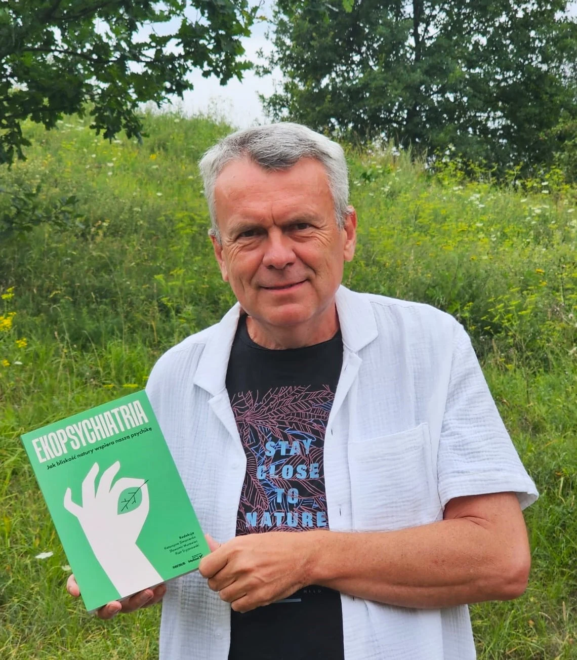 mężczyzna w średnim wieku stoi na tle zielonego wzgórza, ubrany w białą koszulę i czarną koszulkę z napisem 'STAY CLOSE TO NATURE', trzyma w rękach zieloną książkę zatytułowaną 'EKOPSYCHIATRIA' z rysunkiem dłoni i oka na okładce