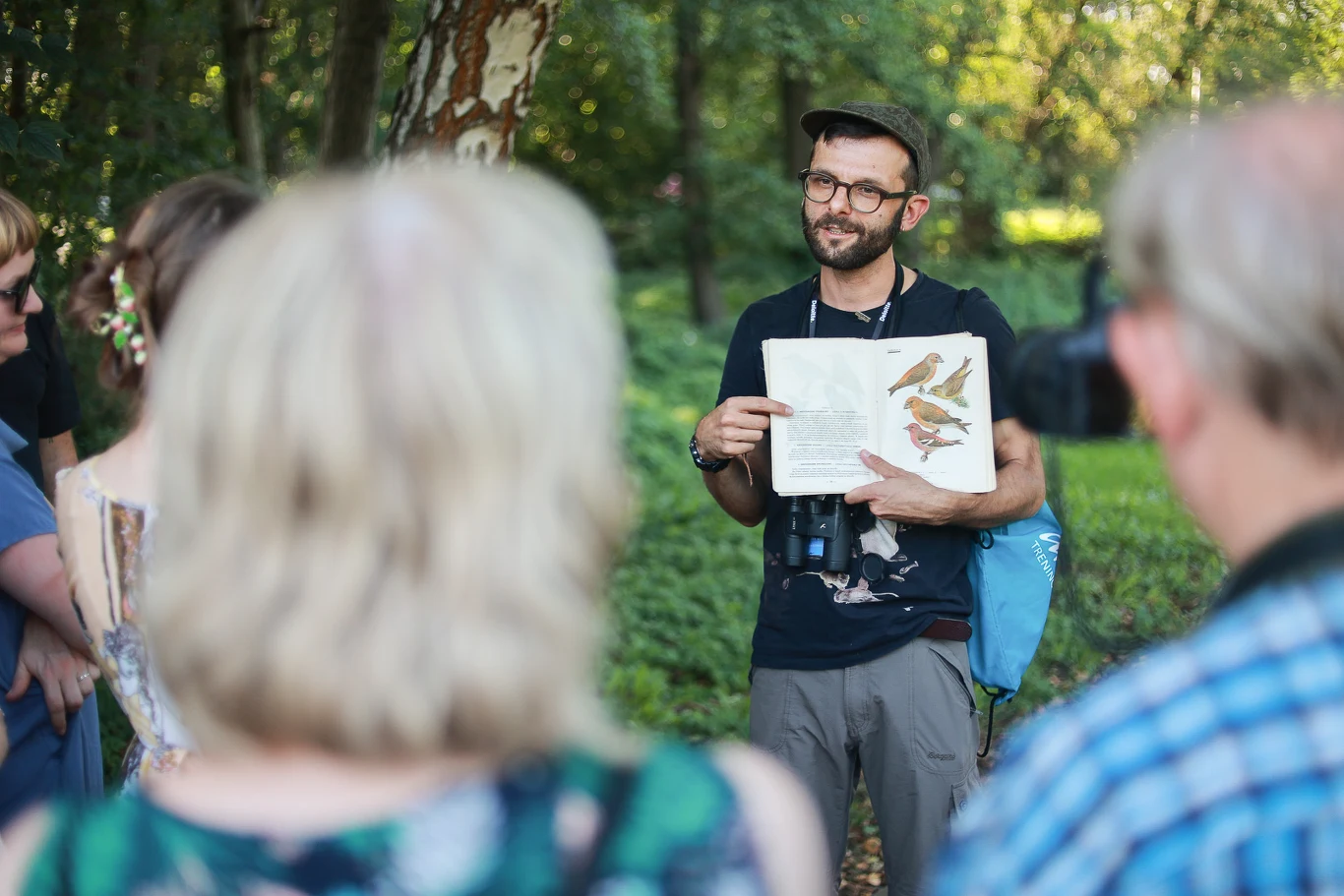 Mężczyzna w okularach i z czapką pokazuje ilustracje ptaków w książce, stojąc w lesie przed grupą słuchaczy; na szyi ma lornetkę, a wokół panuje słoneczny, zielony krajobraz.