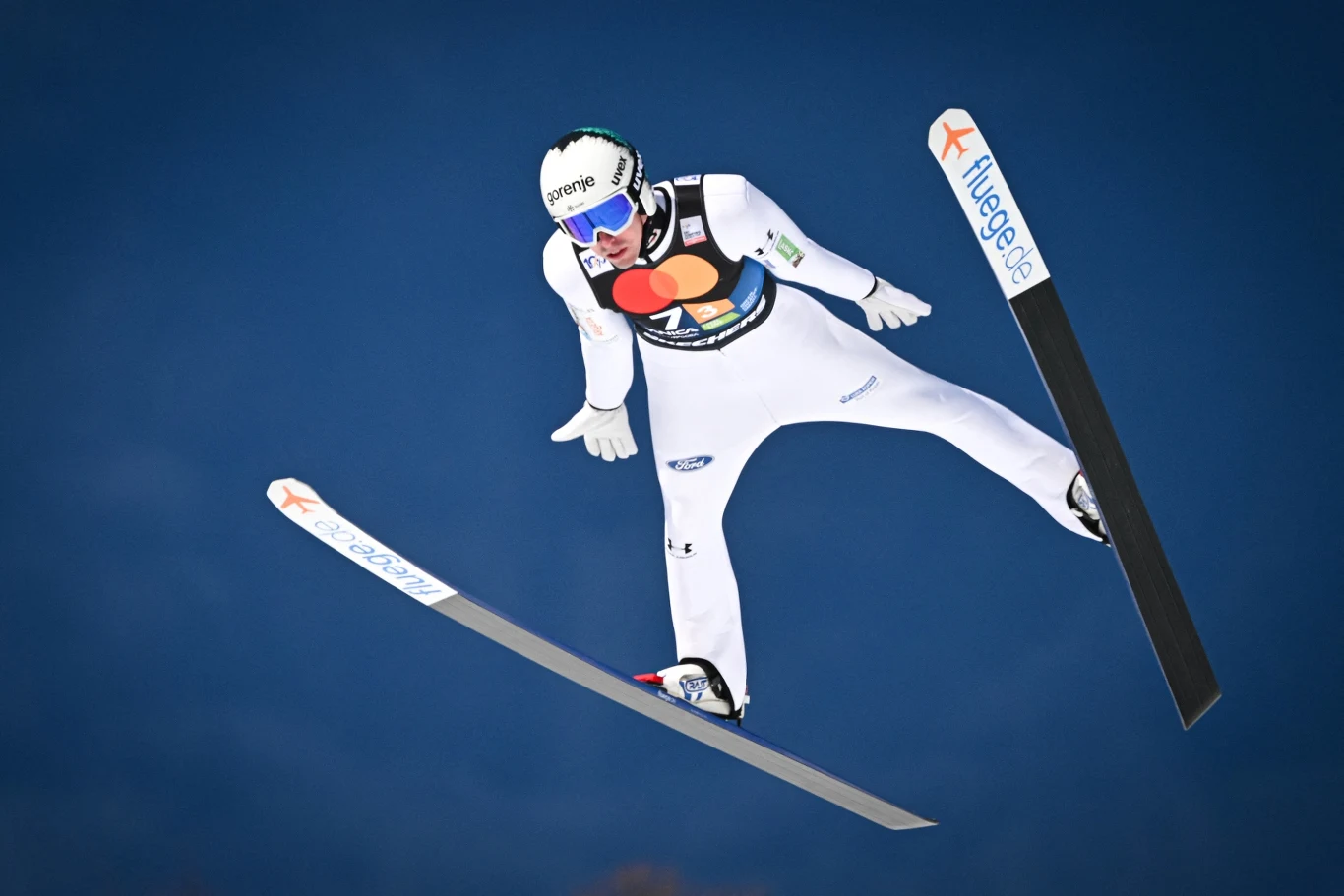 skoczek narciarski w locie z rozłożonymi nartami, ubrany w biały kombinezon sportowy, kask i gogle, dynamiczne ujęcie na tle ciemnego nieba