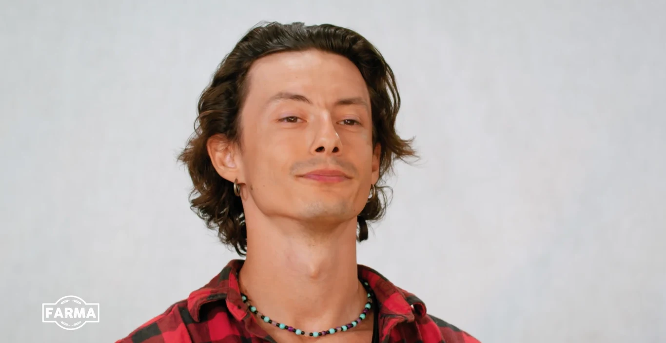 W "Farmie" nie brakuje emocji Mężczyzna z długimi ciemnymi włosami w czerwonej koszuli w kratę i naszyjniku, logo FARMA w lewym dolnym rogu.