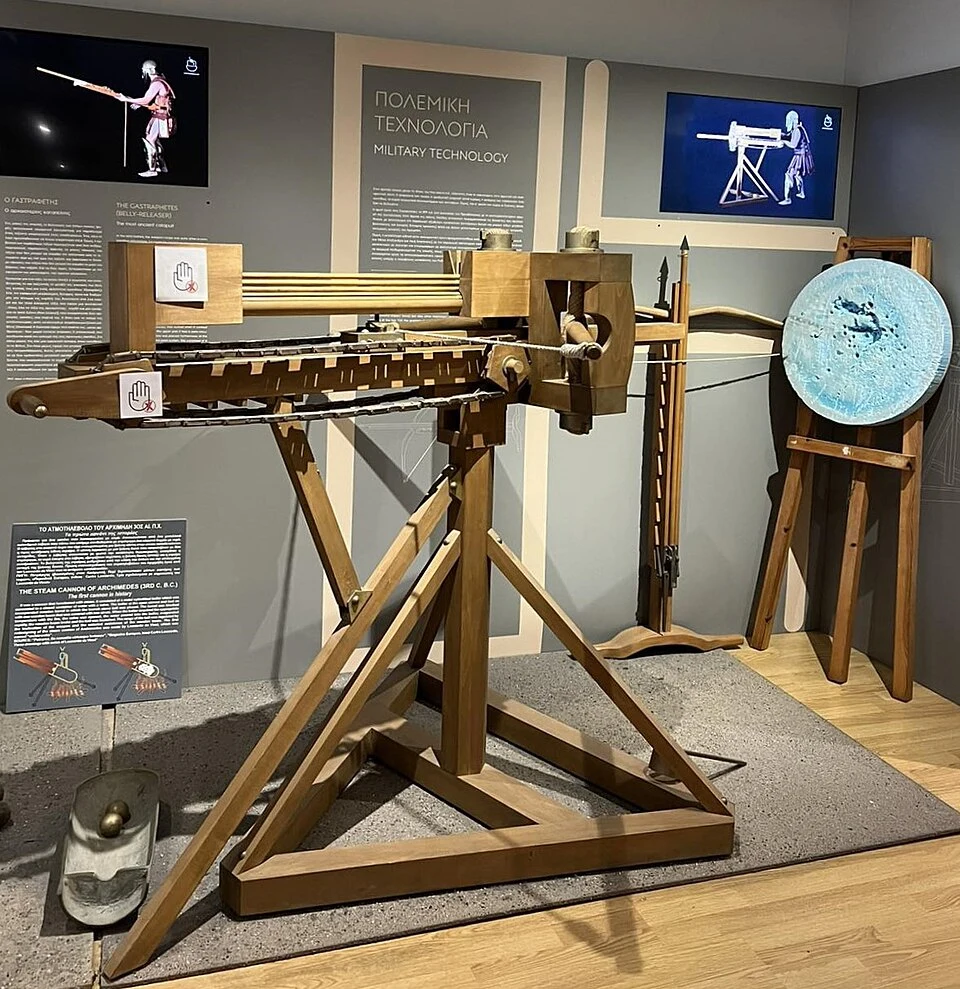 drewniana replika starożytnej machiny oblężniczej typu balista z rozbudowanym mechanizmem naciągu, ustawiona w muzealnej ekspozycji na tle plansz informacyjnych oraz tarczy testowej z widocznymi śladami trafień