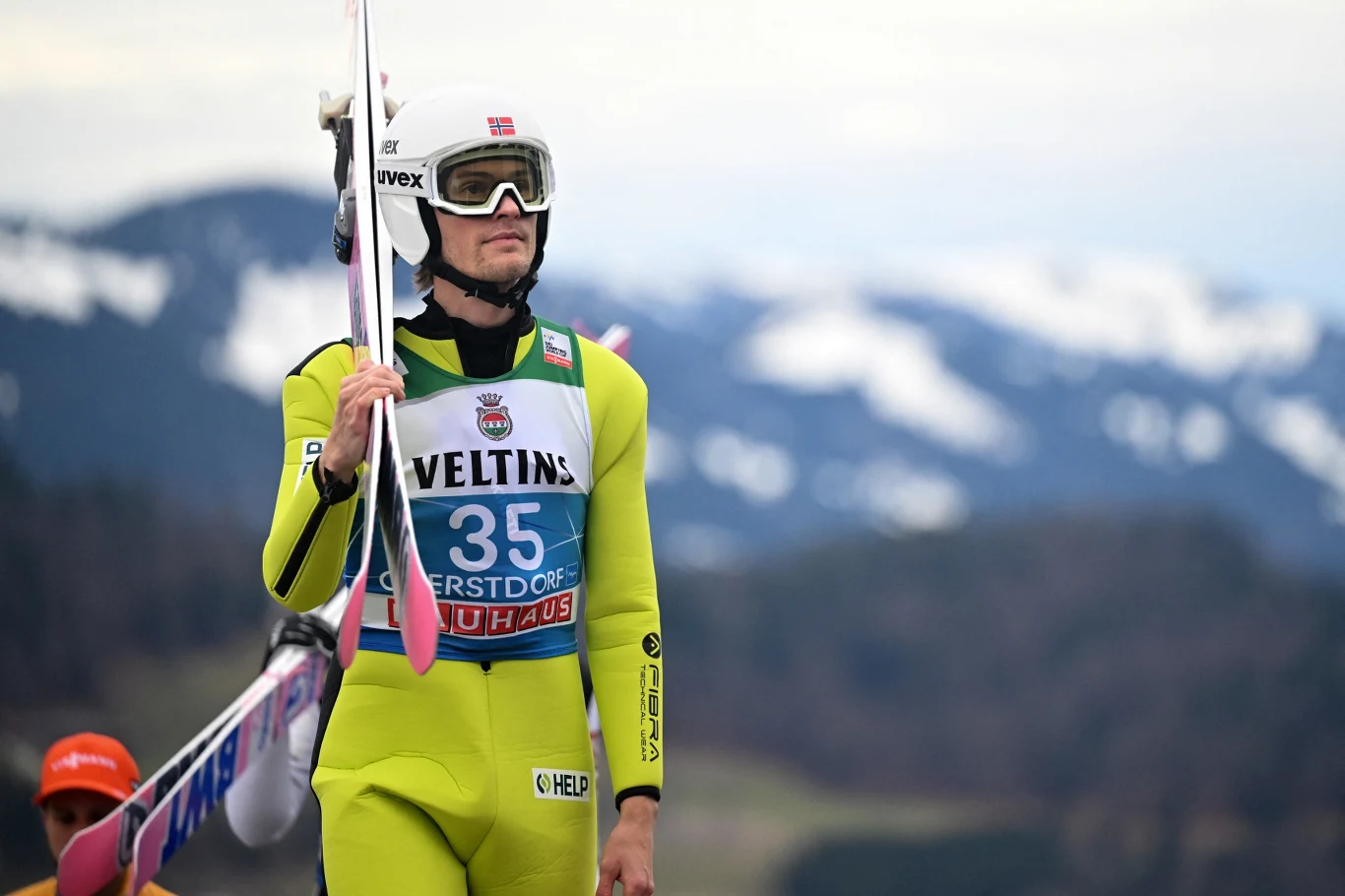 Skoczek narciarski w jasnozielonym kombinezonie i białym kasku trzyma narty na ramieniu; z przodu widoczny numer startowy 35 na tle gór z ośnieżonymi szczytami.