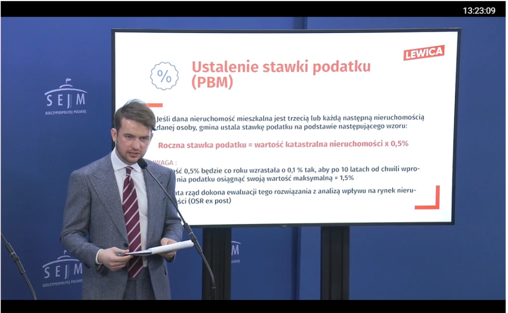 Mężczyzna w garniturze z czerwonym krawatem stoi przy mównicy, obok dużego ekranu prezentującego slajd na temat ustalania stawki podatku od wartości katastralnej nieruchomości, z logotypem 'LEWICA'.