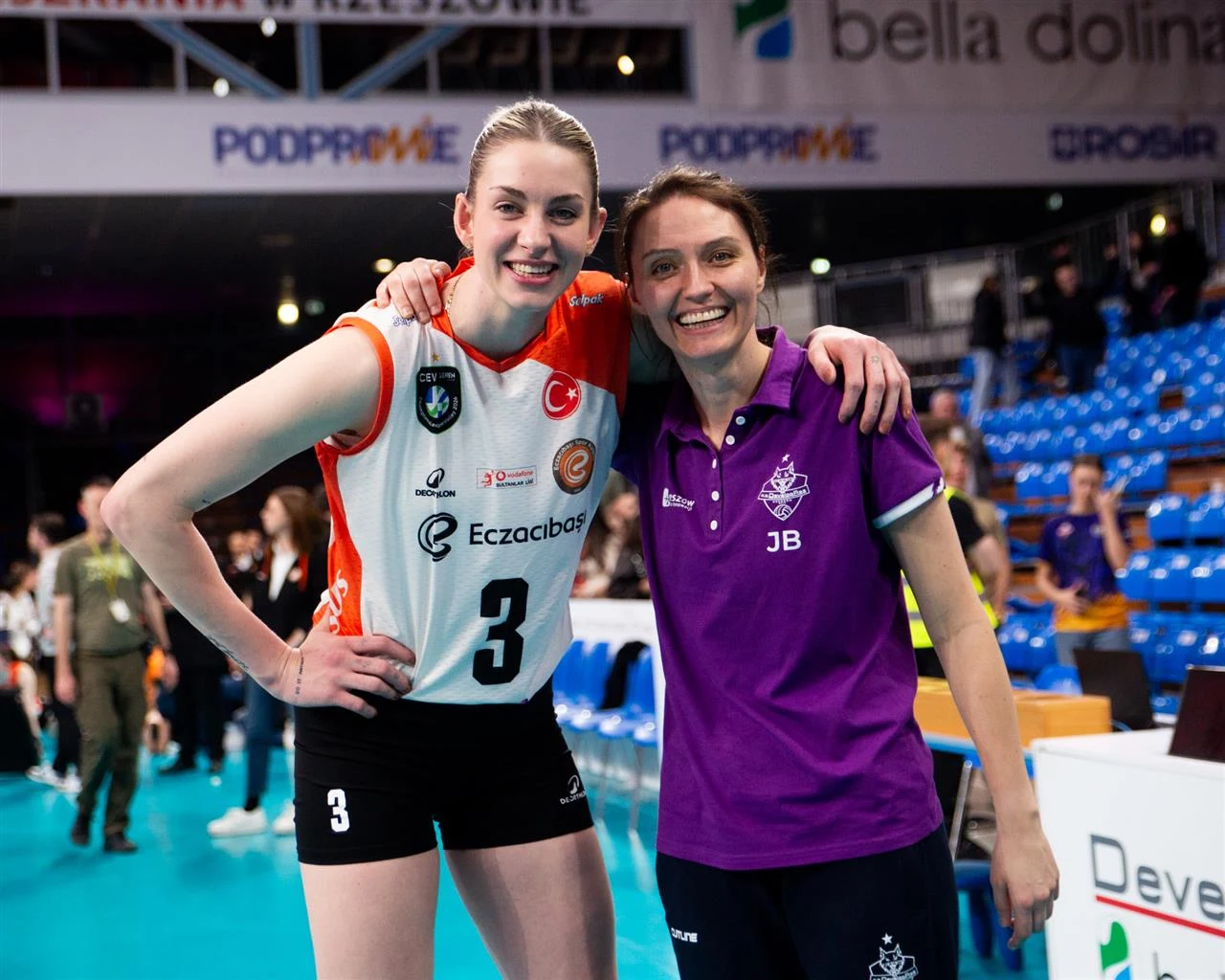 Dwie kobiety na hali sportowej obejmujące się ramionami; jedna ubrana w siatkarską koszulkę z numerem 3, druga w fioletową koszulkę polo z inicjałami JB, obie uśmiechnięte, w tle puste niebieskie trybuny.