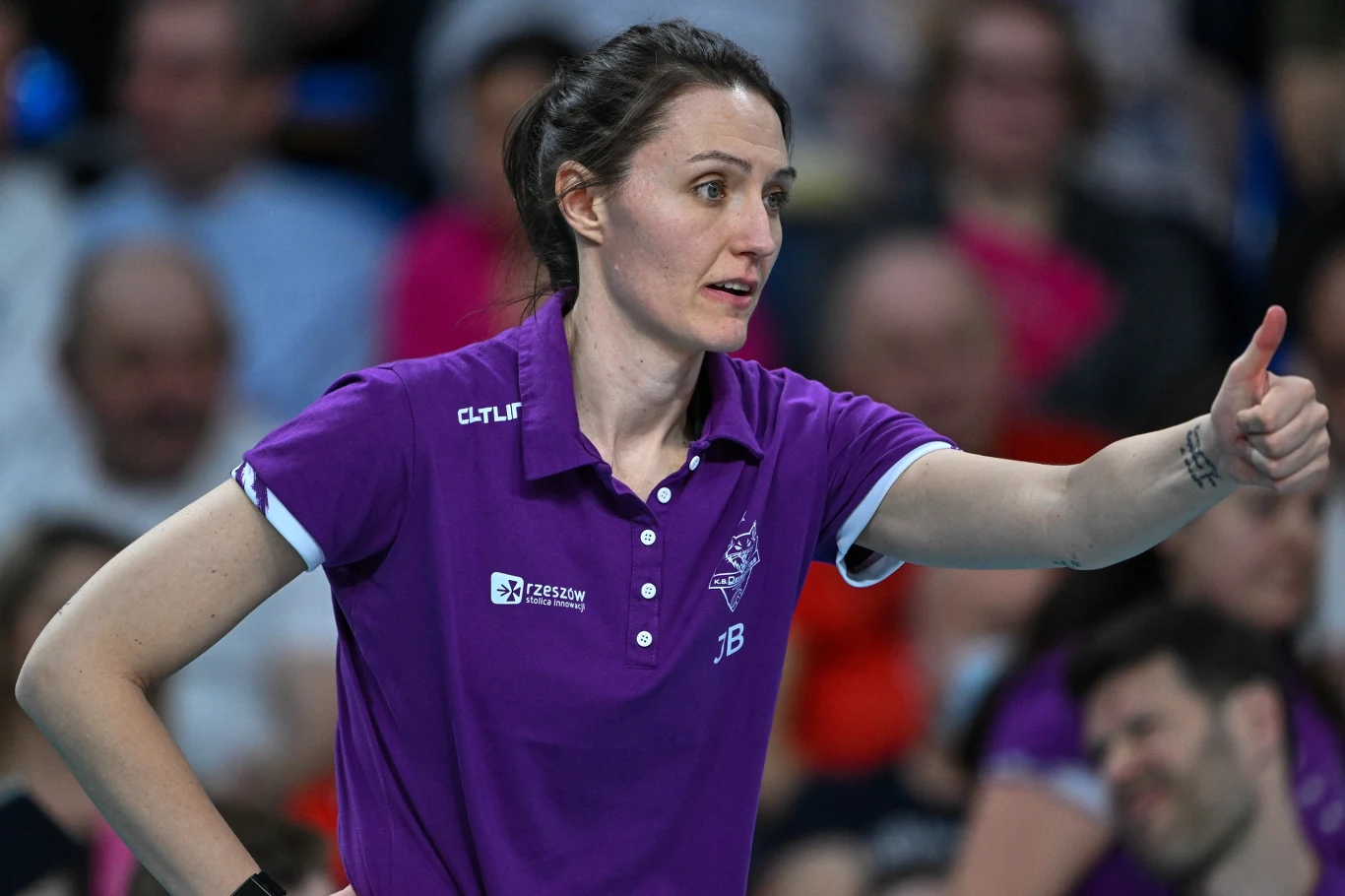 kobieta w fioletowej koszulce sportowej wykonuje gest uniesionego kciuka, sugerujący aprobatę lub zachętę; w tle rozmyta publiczność