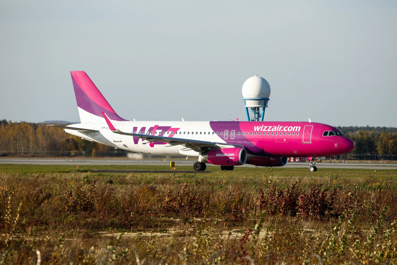Lotnisko w Katowicach Pyrzowicach z roku na rok notuje większy ruch. Samolot Wizz Air kołuje po pasie startowym lotniska Katowice; w tle wieża i jesienne drzewa.