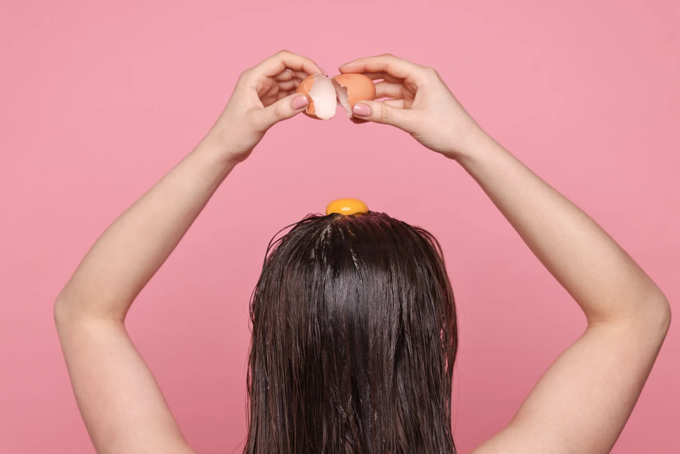 osoba z długimi brązowymi włosami trzyma rozbite jajko nad głową, a żółtko znajduje się na jej włosach, na tle różowej ściany