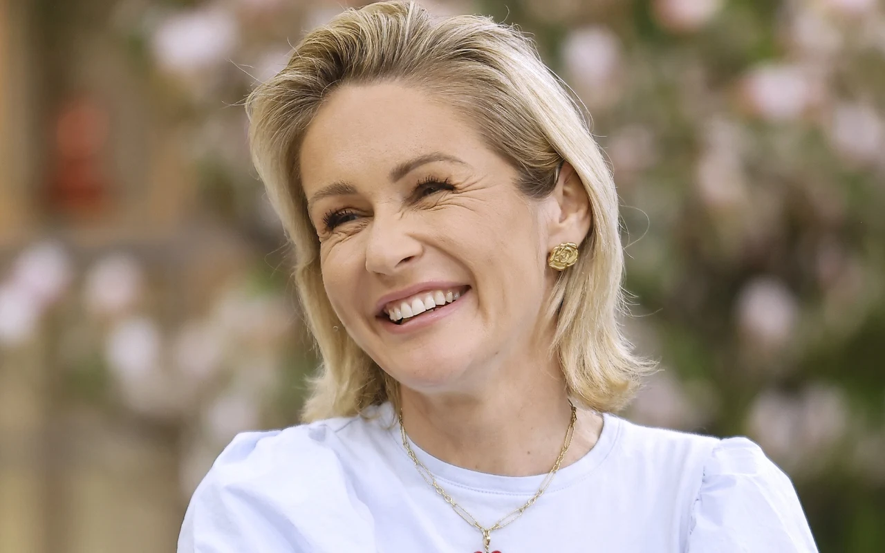 Uśmiechnięta kobieta z blond włosami do ramion, ubrana w jasnoniebieską bluzkę i złote kolczyki, na szyi delikatny łańcuszek z zawieszką; tło rozmyte z różowymi kwiatami.