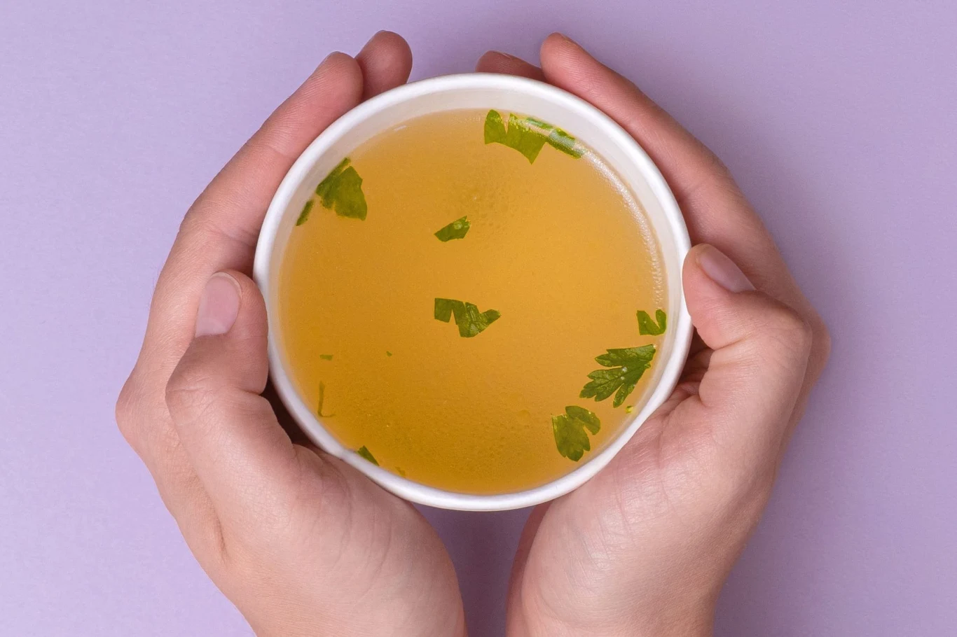 Pyszny i tani bulion z resztek warzyw to świetny wybór Bulion warzywny w białym kubku z kawałkami natki pietruszki, trzymany w dłoniach na fioletowym tle