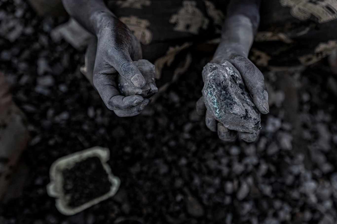 Ręce pokryte pyłem trzymają spory kawałek węgla i mniejszy kamień nad ciemnym tle pełnym węglowych odłamków, w pobliżu plastikowy pojemnik wypełniony drobnymi fragmentami.