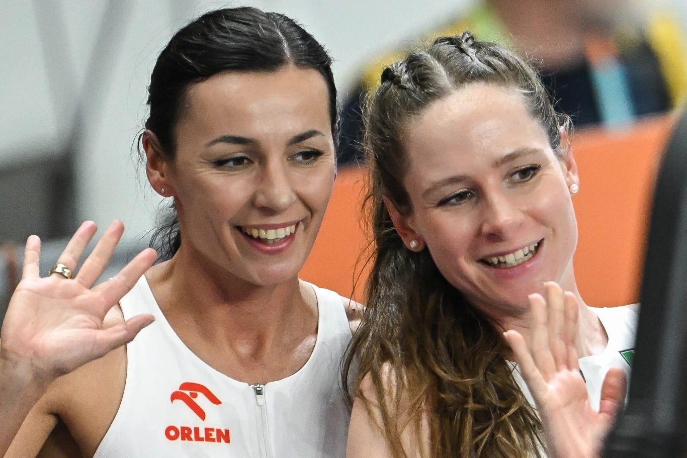 Dwie uśmiechnięte kobiety w sportowych strojach z logotypem ORLEN, machające rękami i patrzące w stronę obiektywu.