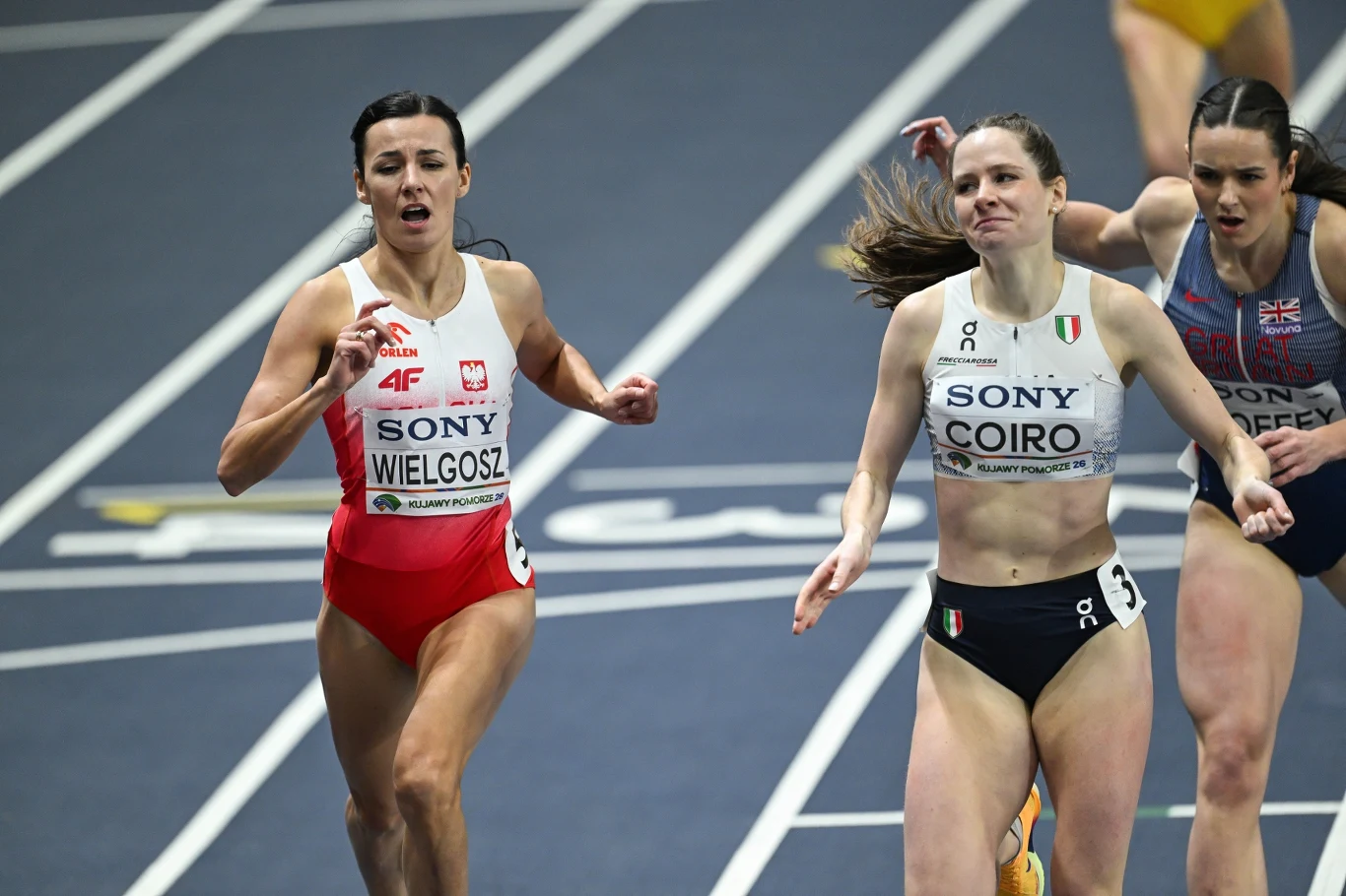 Trzy zawodniczki biegną w wyścigu na hali, skupione na finiszu i rywalizacji o czołowe miejsca, widoczne są stroje sportowe z nazwiskami Wielgosz i Coiro oraz intensywne emocje na twarzach biegaczek.