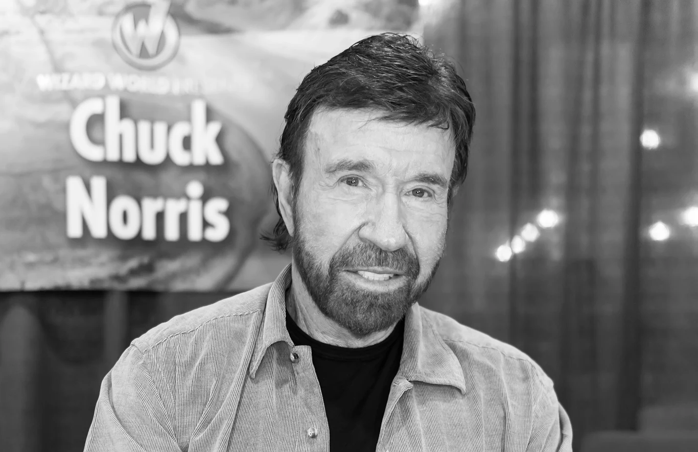 Chuck Norris Chuck Norris, mężczyzna z brodą i krótkimi włosami w koszuli, na tle banera z napisem „Chuck Norris” i ciemnej zasłony.