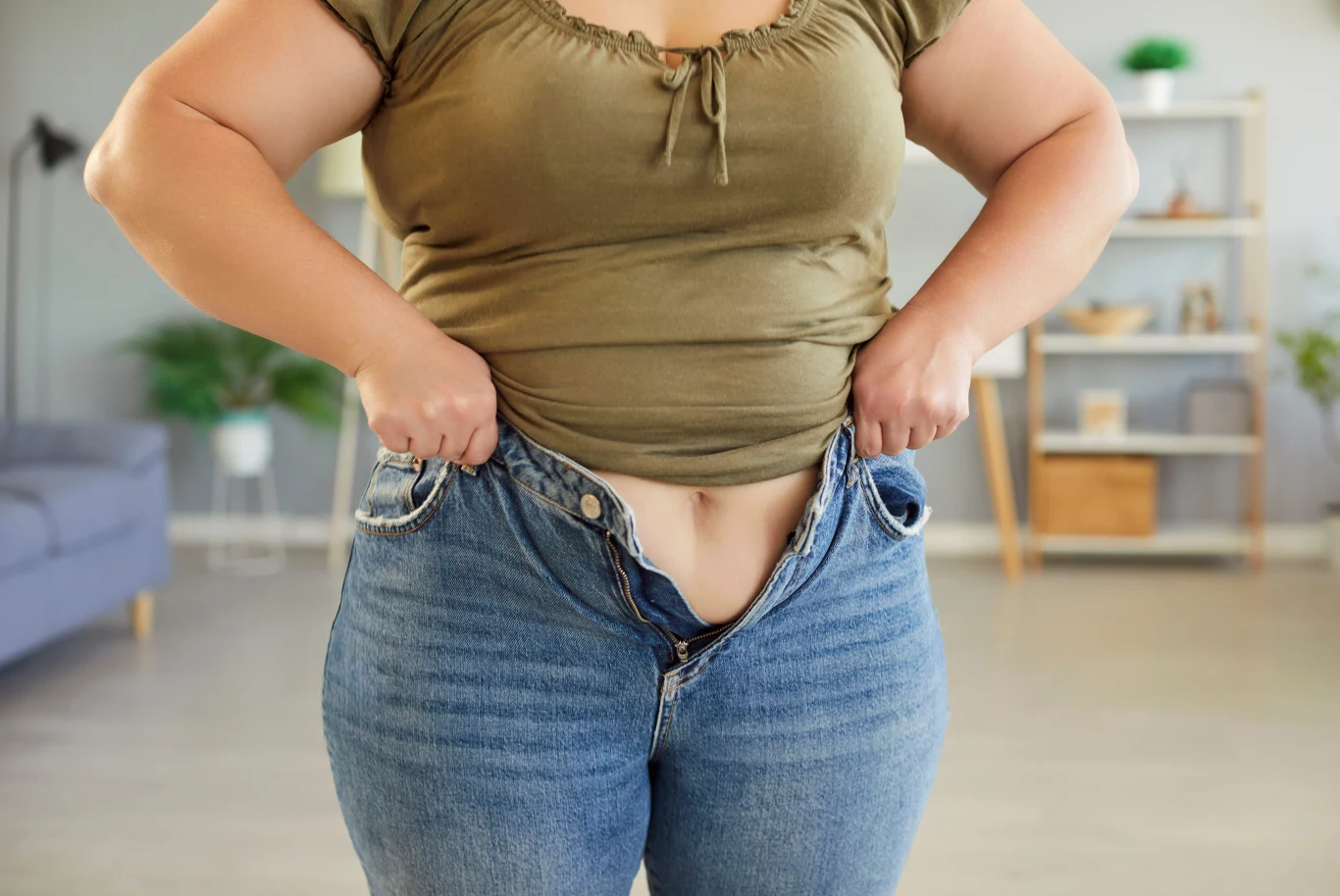 osoba próbująca zapiąć zbyt ciasne dżinsy, ubrana w zieloną koszulkę, znajduje się w jasnym, domowym wnętrzu