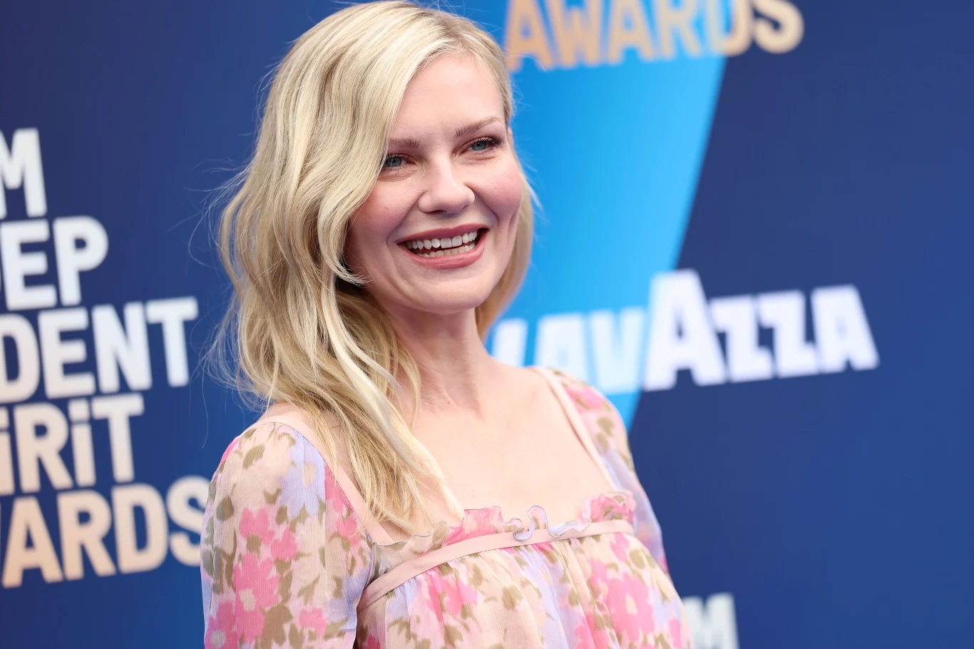 Kobieta z jasnymi, falowanymi włosami uśmiecha się szeroko, ubrana w zwiewną sukienkę w kwiatowy wzór, stojąc na tle niebieskiej ścianki z napisem „INDEPENDENT SPIRIT AWARDS”.