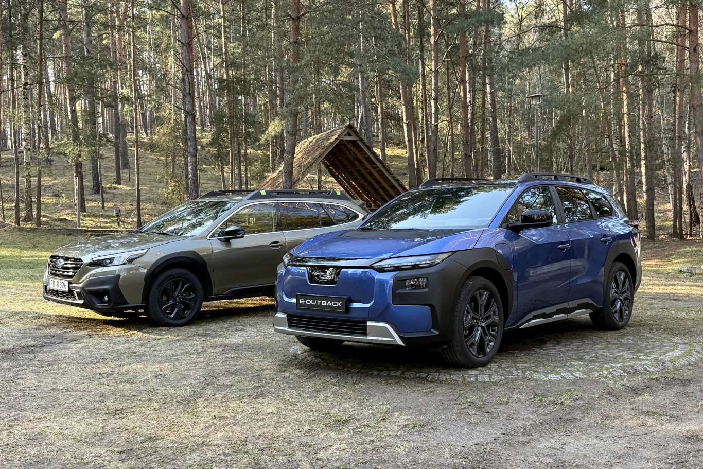 Dwa samochody typu SUV Subaru Outback zaparkowane na leśnej polanie, jeden w kolorze niebieskim, drugi szary, w tle drewniana wiata i wysokie sosny.