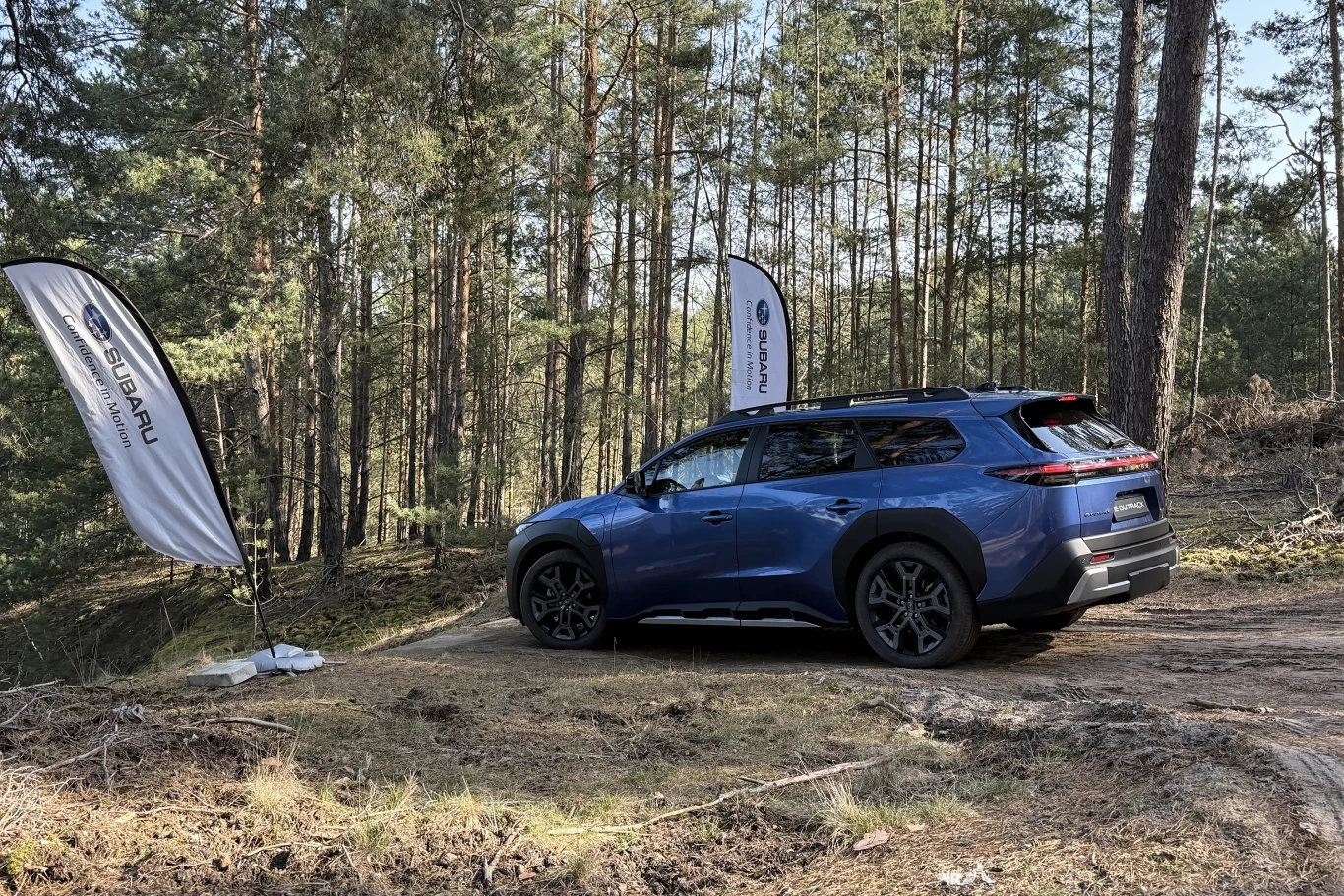 niebieski SUV Subaru zaparkowany na leśnej drodze, otoczony sosnami, obok pojazdu stoją dwie flagi z logo Subaru