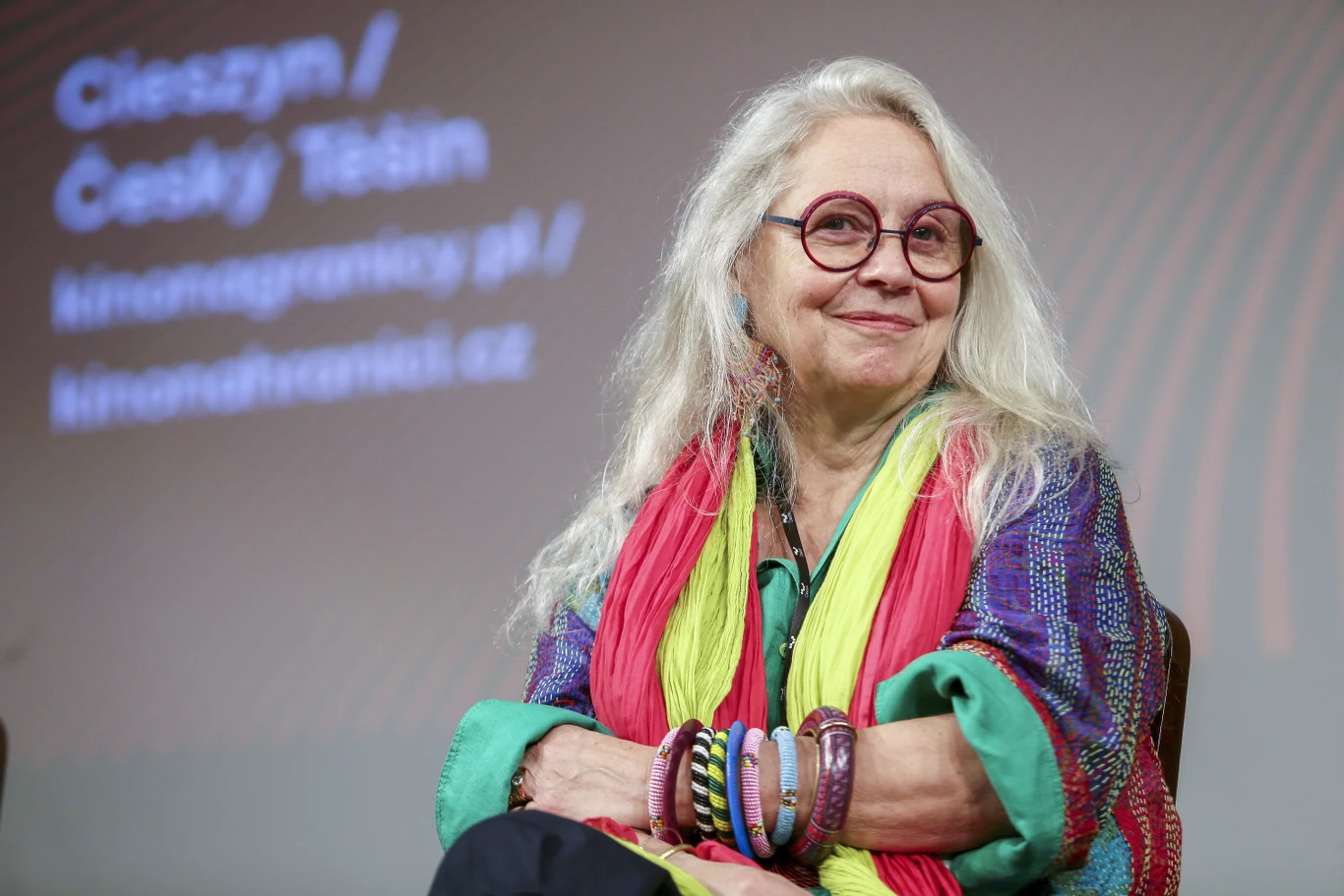 starsza kobieta z długimi siwymi włosami i okrągłymi okularami siedzi uśmiechnięta, ubrana w kolorowe, wzorzyste ubrania oraz liczne bransoletki, w tle rozmazany napis z nazwami miejscowości i adresami stron internetowych