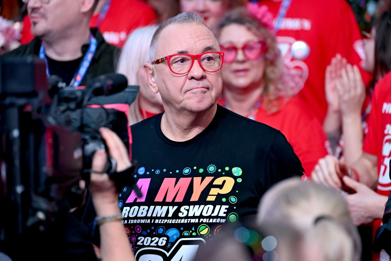 Jurek Owsiak stoi na czele Pol'and'Rock Festivalu Jurek Owsiak w czarnej koszulce i czerwonych okularach wśród osób w czerwonych koszulkach, przed kamerą TV