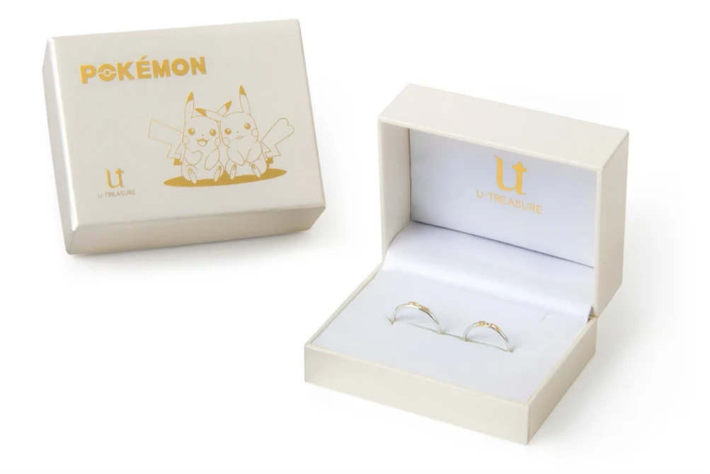 białe pudełko z wizerunkiem dwóch Pikachu oraz napisem Pokémon, obok otwarte eleganckie etui z dwoma złotymi pierścionkami na białym welurowym tle