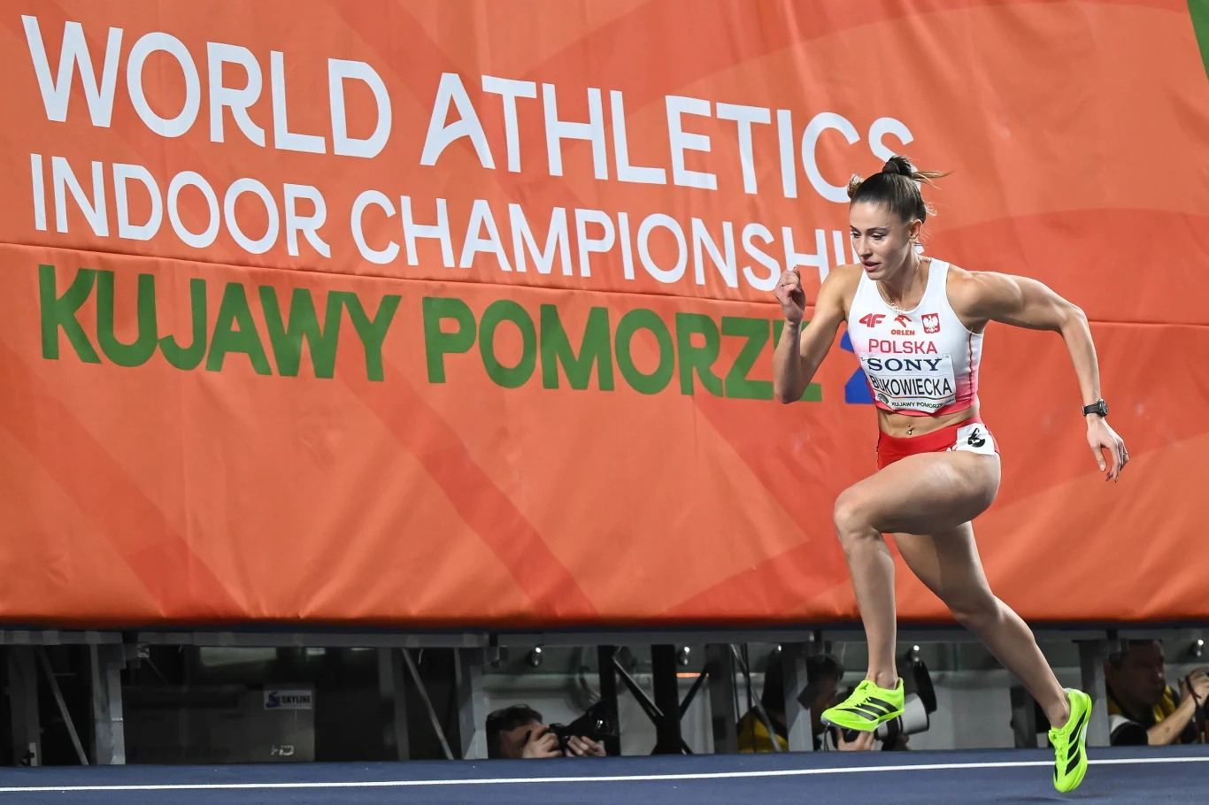 Lekkoatletka z Polski w stroju reprezentacyjnym biegnie po bieżni podczas halowych mistrzostw świata, dynamiczny ruch ciała podkreślony przez zaciśnięte pięści i skoncentrowane spojrzenie, w tle widoczne duże logo imprezy na pomarańczowym banerze.