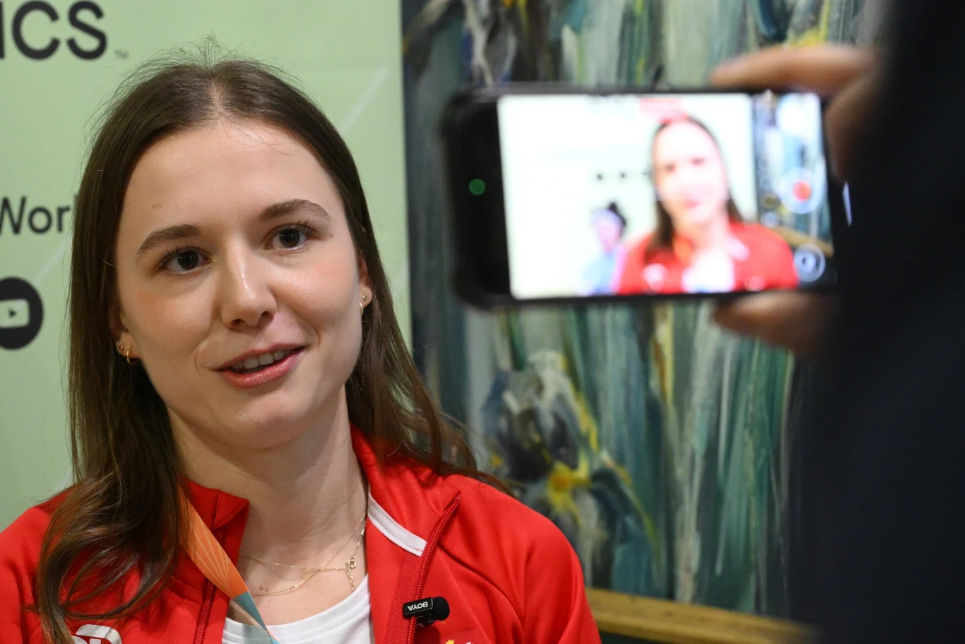 Młoda kobieta w czerwonej marynarce udziela wywiadu, mając przypięty mikrofon do ubrania, a jej obraz jest nagrywany smartfonem trzymanym przez inną osobę.