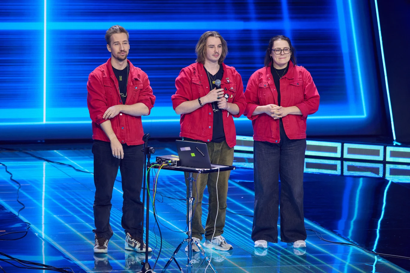 Zespół Imasleep w "Must Be The Music" Trójka osób ubranych w czerwone kurtki stoi na scenie oświetlonej niebieskimi światłami, przed nimi znajduje się mały stolik z laptopem i kablami.