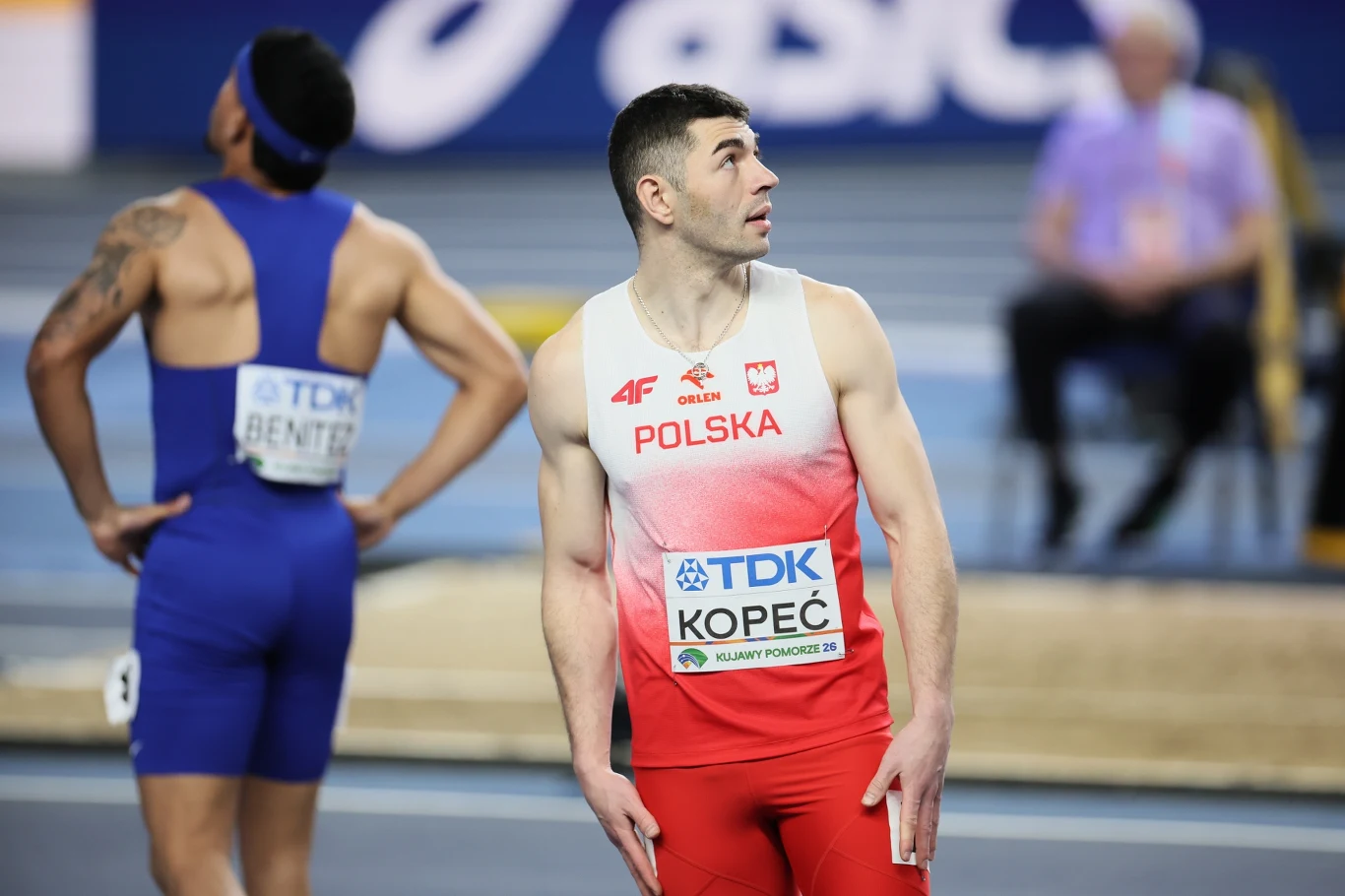 sprinter w biało-czerwonym stroju z napisem POLSKA i nazwiskiem Kopeć na plakietce spoglądający w bok na bieżni lekkoatletycznej, obok niego inny zawodnik w niebieskim stroju z rękoma opartymi na biodrach