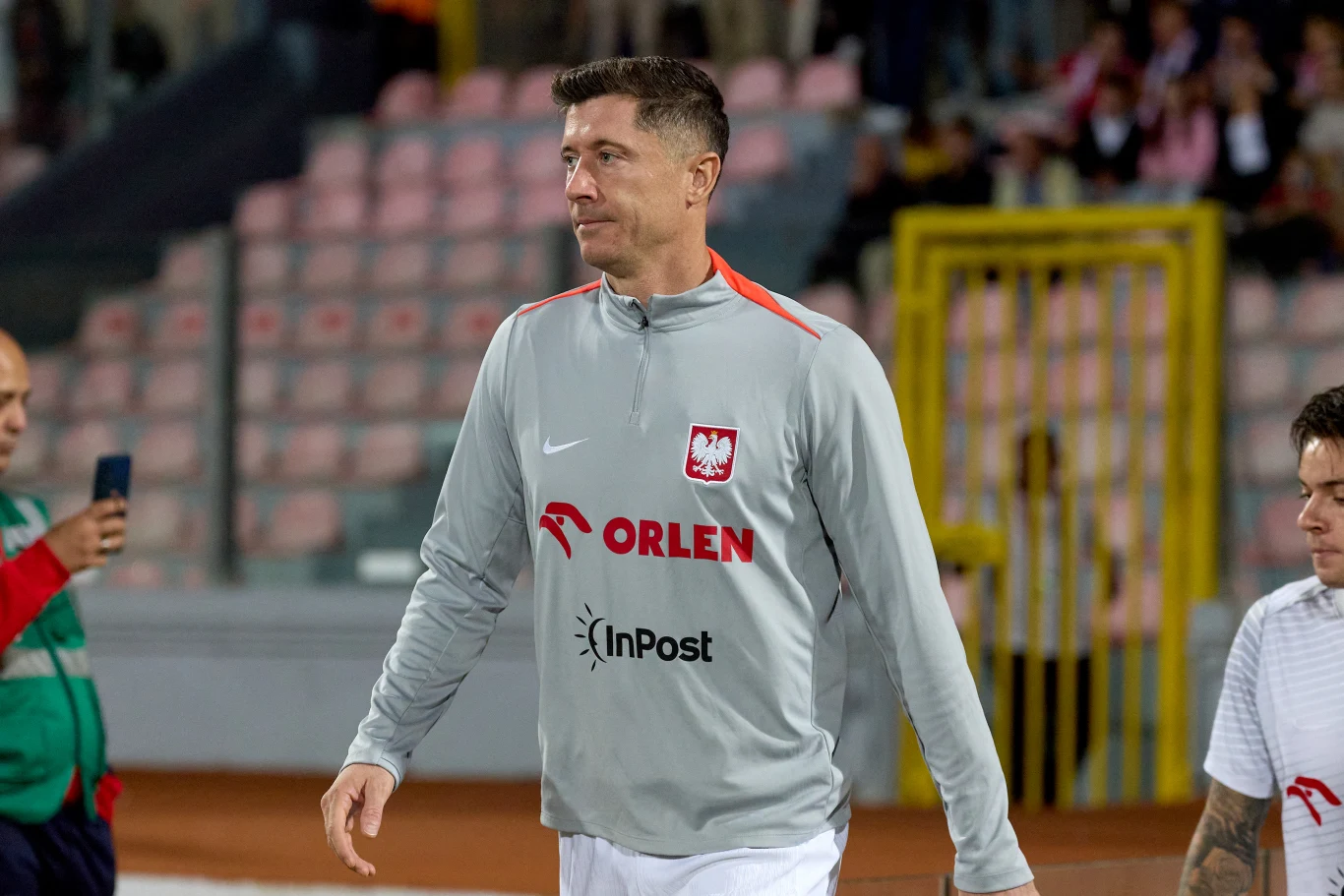 piłkarz ubrany w szary dres z godłem Polski oraz logotypami sponsorów, idący po murawie stadionu podczas przygotowań do meczu