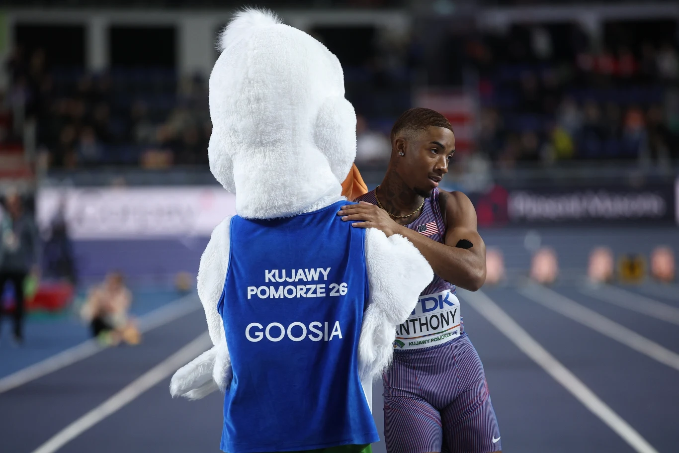 zawodnik lekkoatletyczny w sportowym stroju przytula maskotkę w niebieskiej kamizelce z napisem 'KUJAWY POMORZE 26 GOOSIA' na torze stadionu lekkoatletycznego, w tle niewyraźni kibice i charakterystyczne wyznaczone linie bieżni