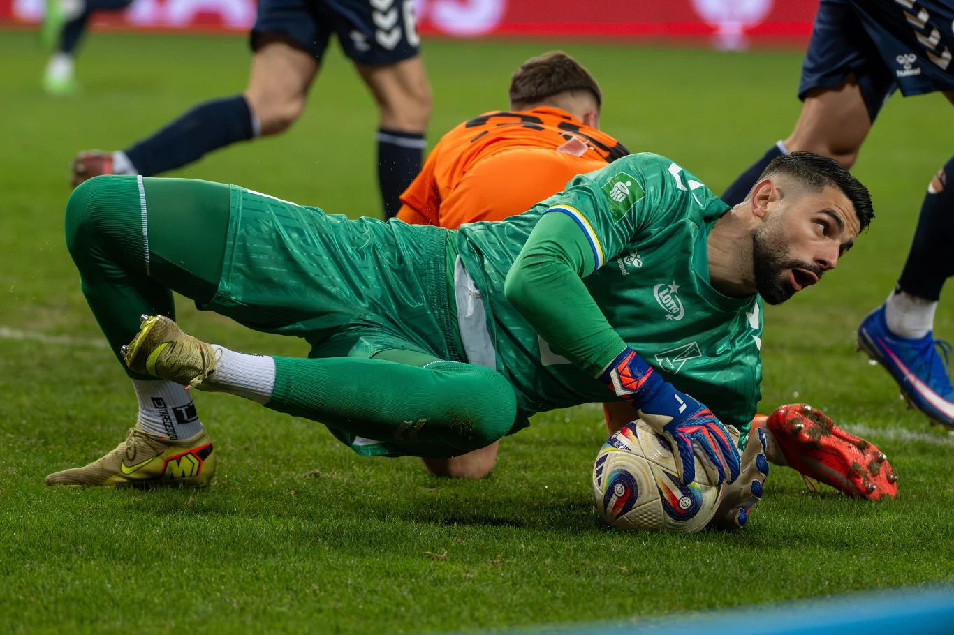 bramkarz w zielonym stroju łapie piłkę na murawie stadionu, wokół niego znajdują się zawodnicy próbujący przejąć futbolówkę