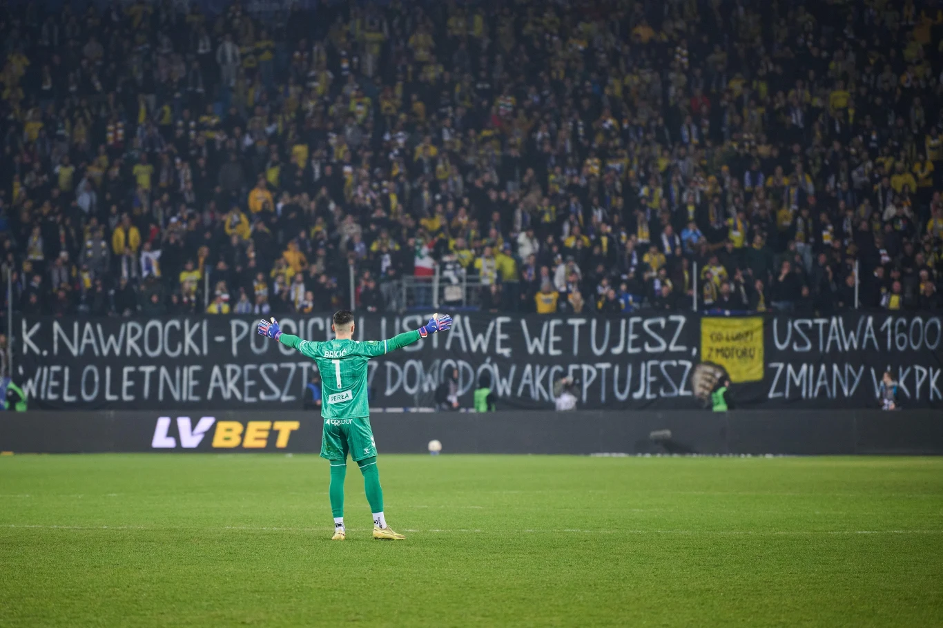 Bramkarz w zielonym stroju stoi tyłem na murawie stadionu z uniesionymi rękami, w tle tłum kibiców z żółto-czarnymi akcesoriami oraz duży transparent z hasłami skierowanymi do konkretnej osoby.