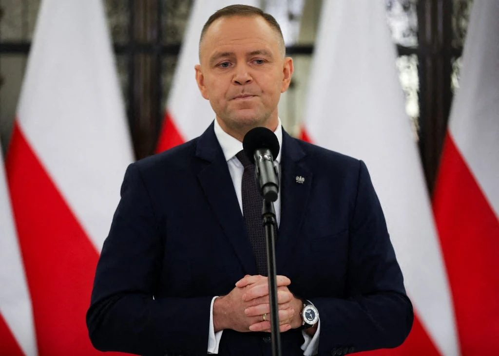 Mężczyzna w garniturze stoi za mikrofonem na tle kilku polskich flag, trzymając dłonie splecione przed sobą, sprawiając wrażenie powagi i skupienia.