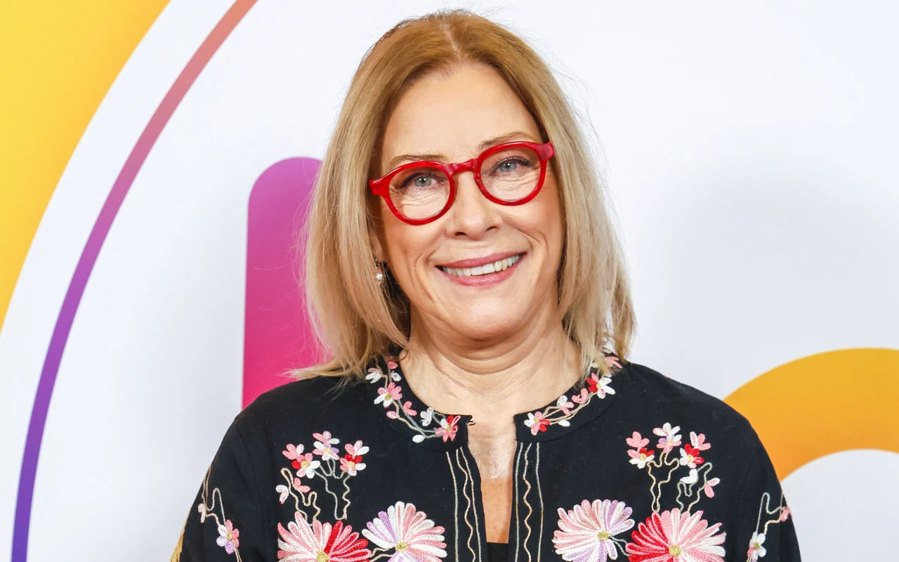 Kobieta w średnim wieku z blond włosami nosi czerwone okulary i czarną bluzkę ozdobioną kolorowymi, kwiatowymi haftami; stoi na tle z kolorowymi łukami i jasnym światłem.