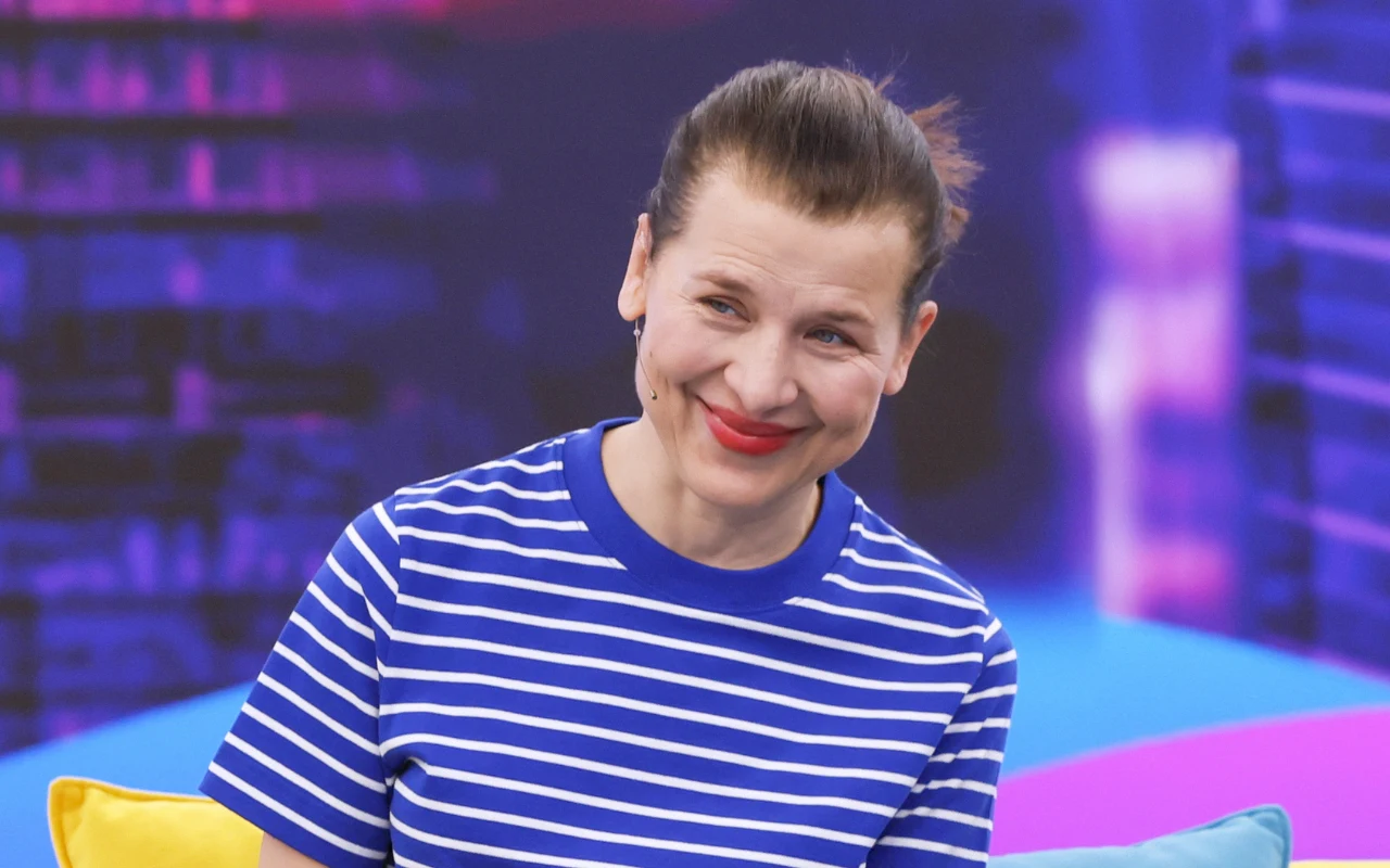 Uśmiechnięta kobieta w pasiastym, niebiesko-białym t-shircie z włosami spiętymi do tyłu, siedzi na kanapie z żółtą i błękitną poduszką, na tle kolorowego, rozmytego tła.