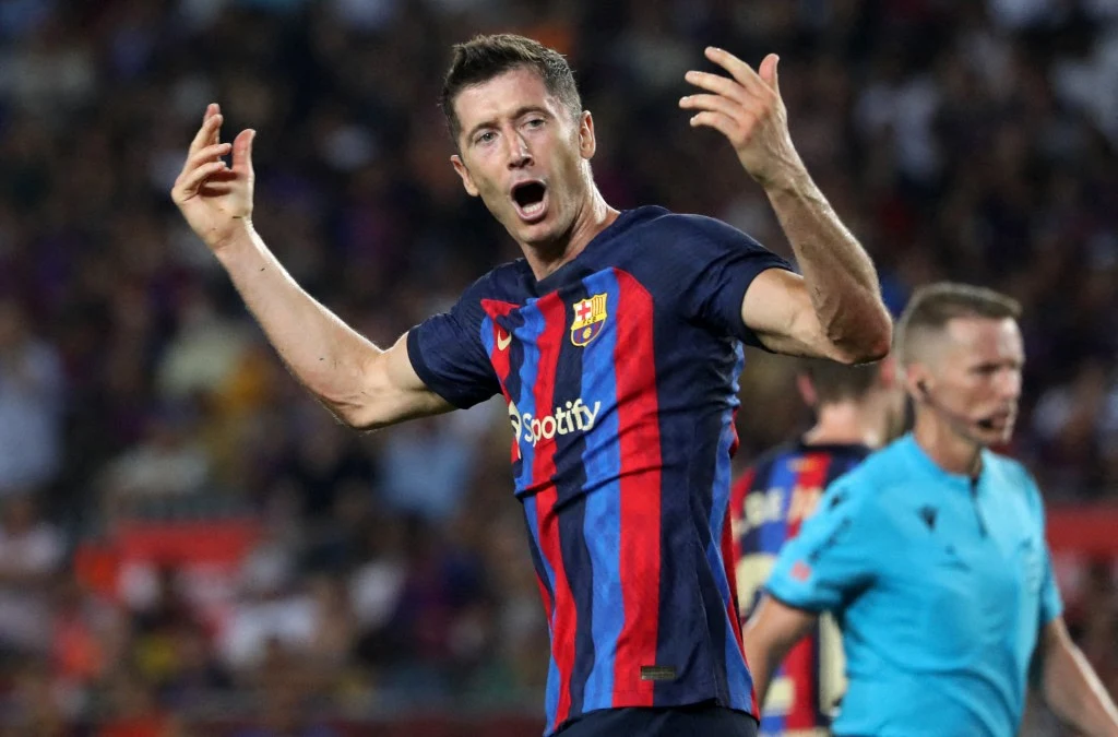 piłkarz w koszulce FC Barcelony z rozłożonymi ramionami i otwartymi ustami, w tle sędzia oraz rozmyty tłum kibiców