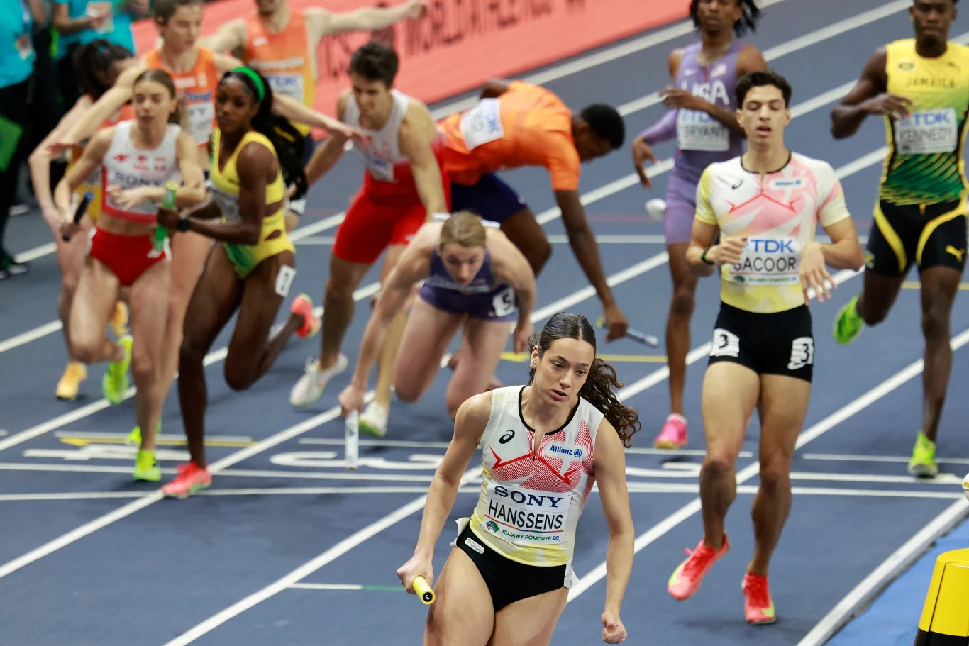 Zawodniczka z pałeczką sztafetową biegnie na prowadzeniu na hali lekkoatletycznej, za nią widoczni pozostali rywale, wśród których niektórzy przewracają się podczas przekazywania pałeczki.