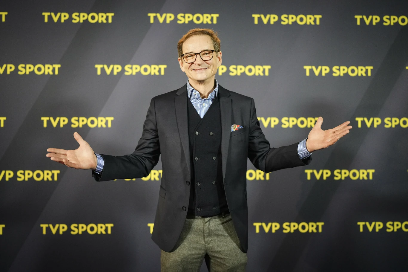 Uśmiechnięty mężczyzna w okularach, ubrany elegancko w marynarkę, kamizelkę i koszulę, stoi z szeroko rozłożonymi rękami na tle ścianki z logo TVP Sport.