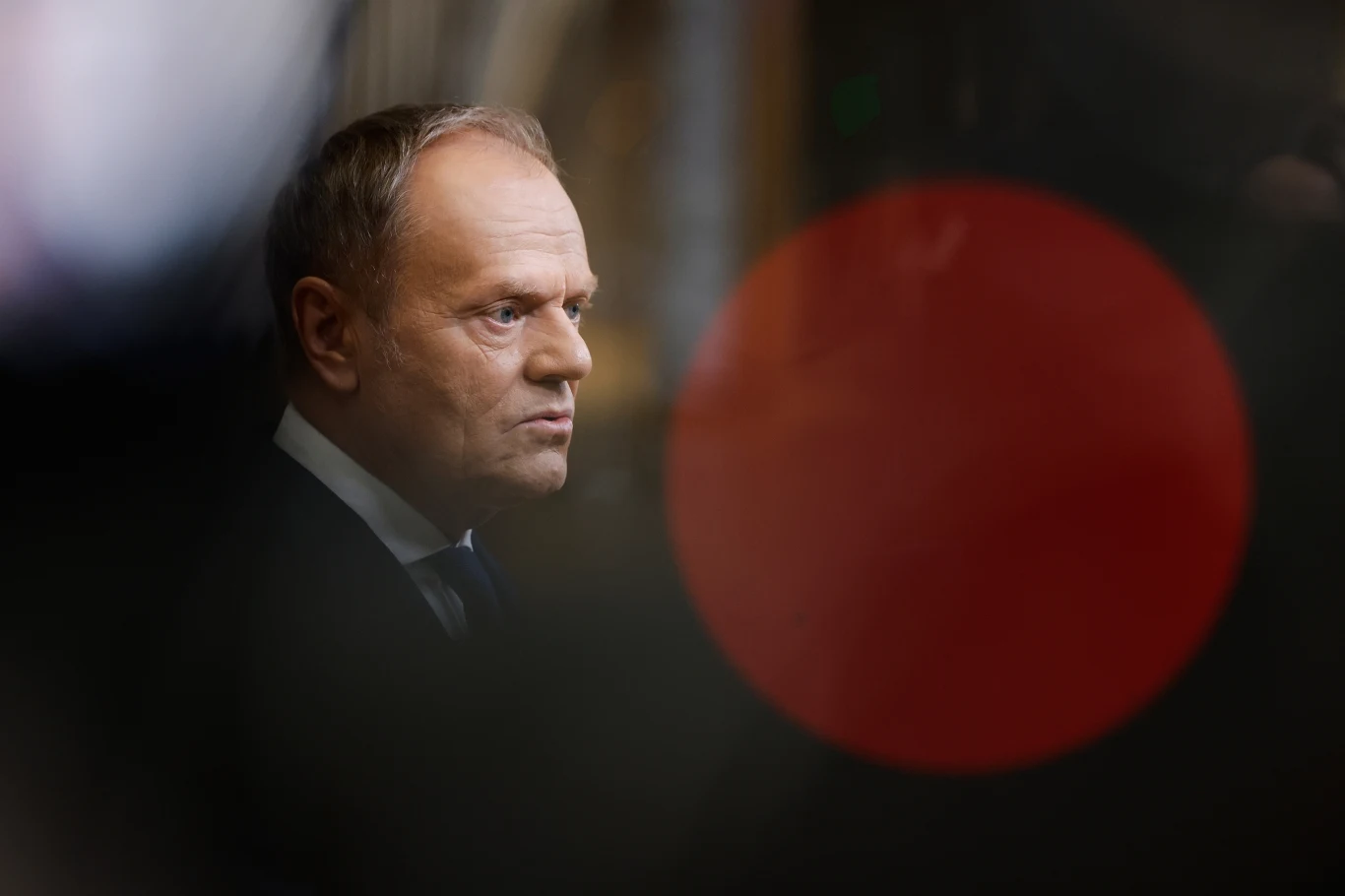 Premier Polski Donald Tusk Mężczyzna w garniturze i podświetlonym tle, na pierwszym planie duże, rozmazane czerwone koło przykuwające uwagę.