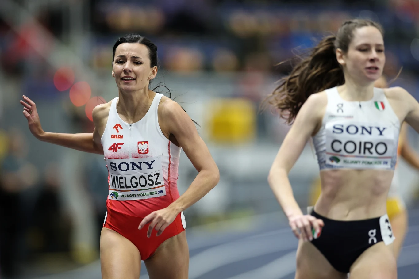 dwie zawodniczki biegną przez linię mety podczas zawodów lekkoatletycznych, obie skupione na finiszu, jedna w biało-czerwonym stroju z orłem i nazwiskiem Wielgosz, druga w biało-czarnym stroju z nazwiskiem Coiro, dynamiczny moment rywalizacji