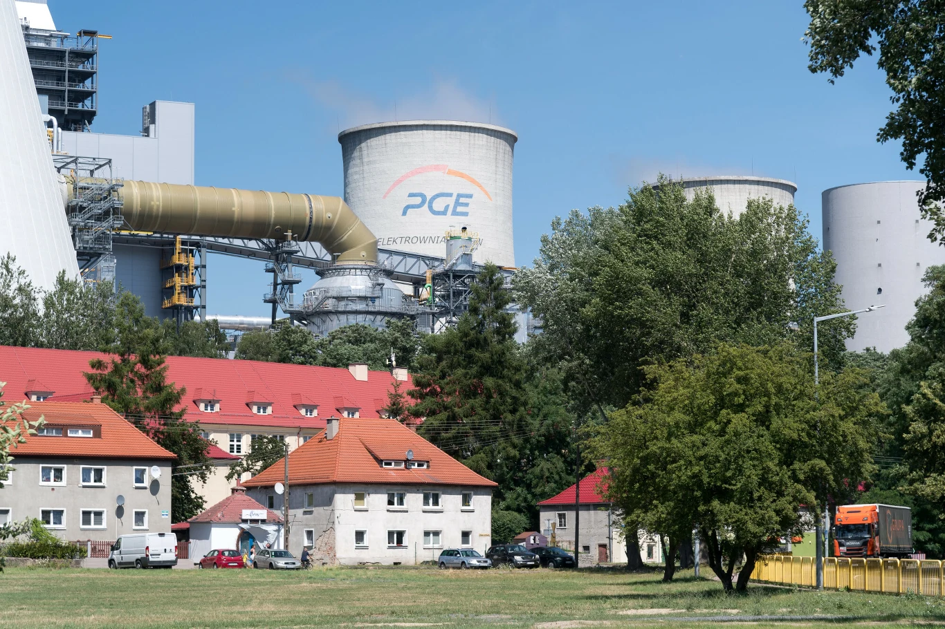 wysokie chłodnie kominowe i przemysłowe instalacje elektrowni PGE dominujące nad osiedlem niskich bloków mieszkalnych z czerwonymi dachami, otoczonych zielenią i drzewami; na ulicy stoi pomarańczowa ciężarówka
