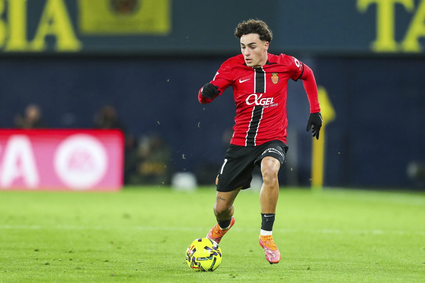 Piłkarz w czerwono-czarnej koszulce z numerem 17 prowadzi piłkę po zielonym boisku podczas meczu piłkarskiego.