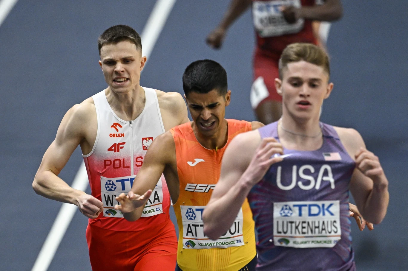 Trzech lekkoatletów biegnie na różnokolorowych torach stadionu, każdy w stroju reprezentującym swój kraj: z Polski, Hiszpanii i USA, skupieni na finiszu biegu, z widocznymi napięciem i wysiłkiem na twarzach.