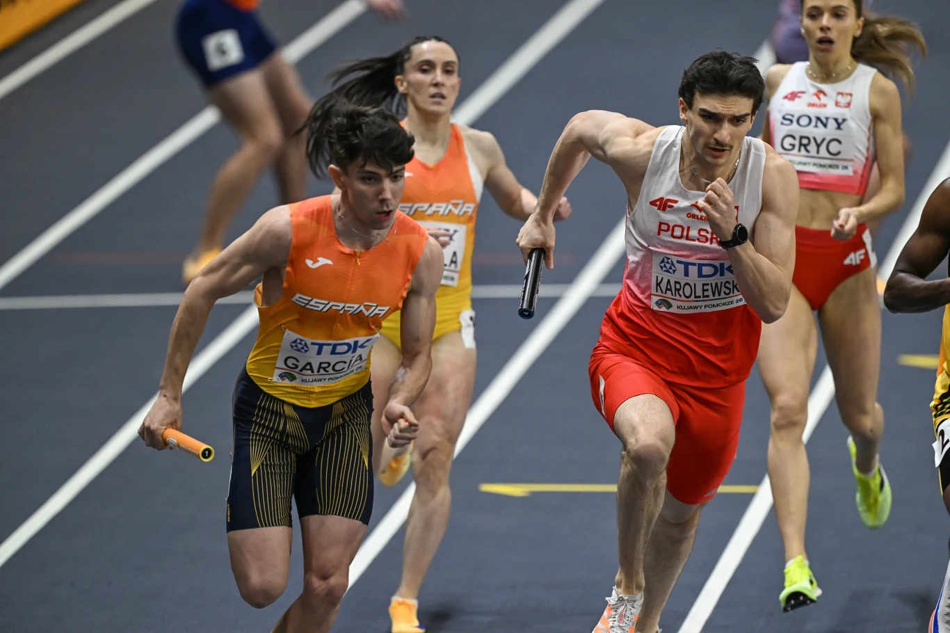 Zawodnicy z różnych krajów biegną w sztafecie na hali, każdy trzyma pałeczkę, widoczne napięcie i skupienie na twarzach, wyróżniają się reprezentacyjne stroje i dynamiczny ruch.