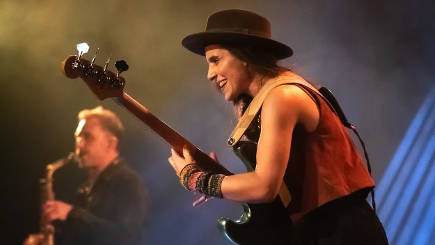 kobieta w kapeluszu grająca na gitarze basowej na scenie, w tle saksofonista, ciepłe światło sceniczne podkreślające ekspresję i energię występu