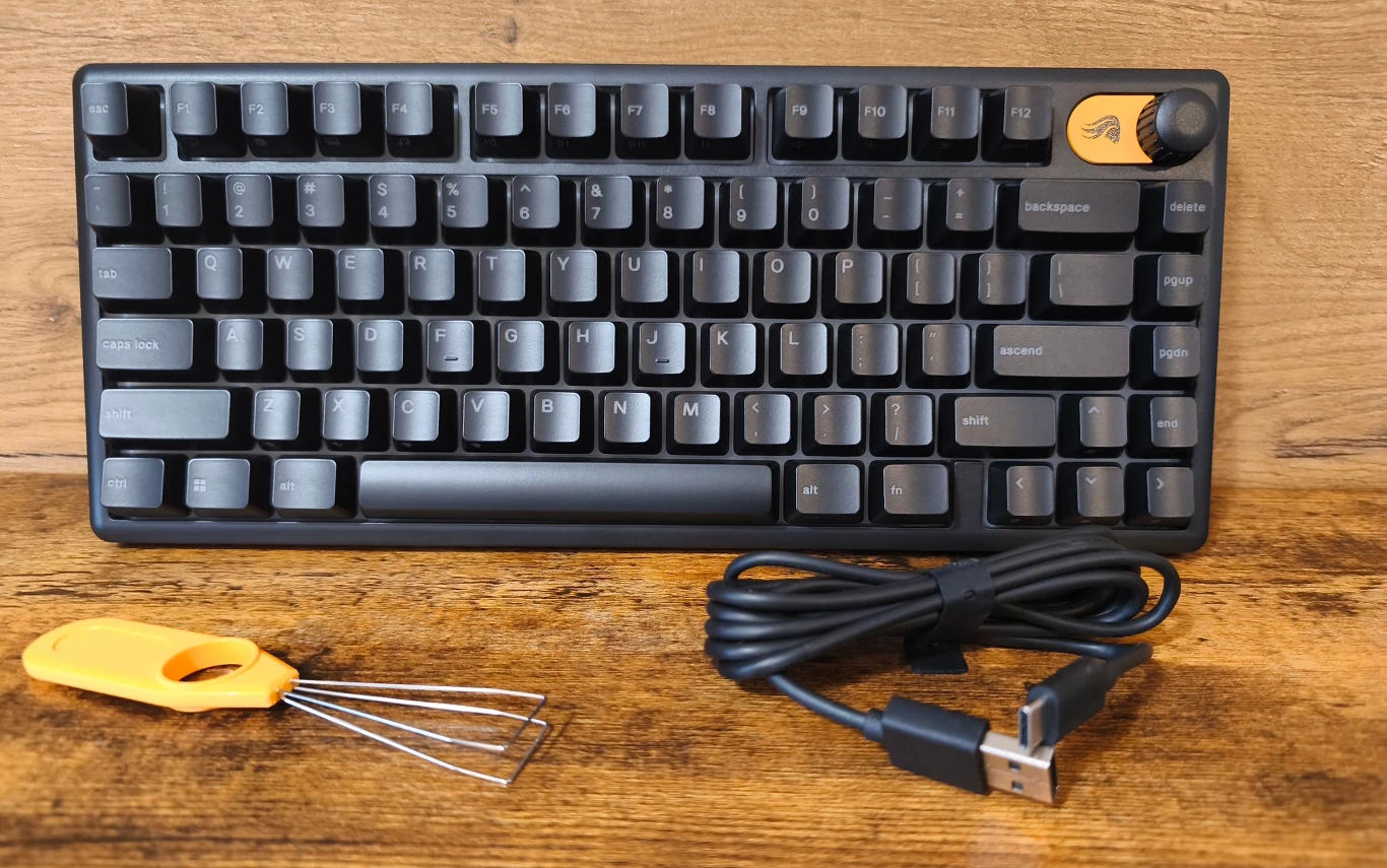 Czarna klawiatura mechaniczna z żółtym przyciskiem funkcyjnym w prawym górnym rogu i pokrętłem, obok leży czarny kabel USB oraz narzędzie do wyciągania klawiszy z żółtą rączką.